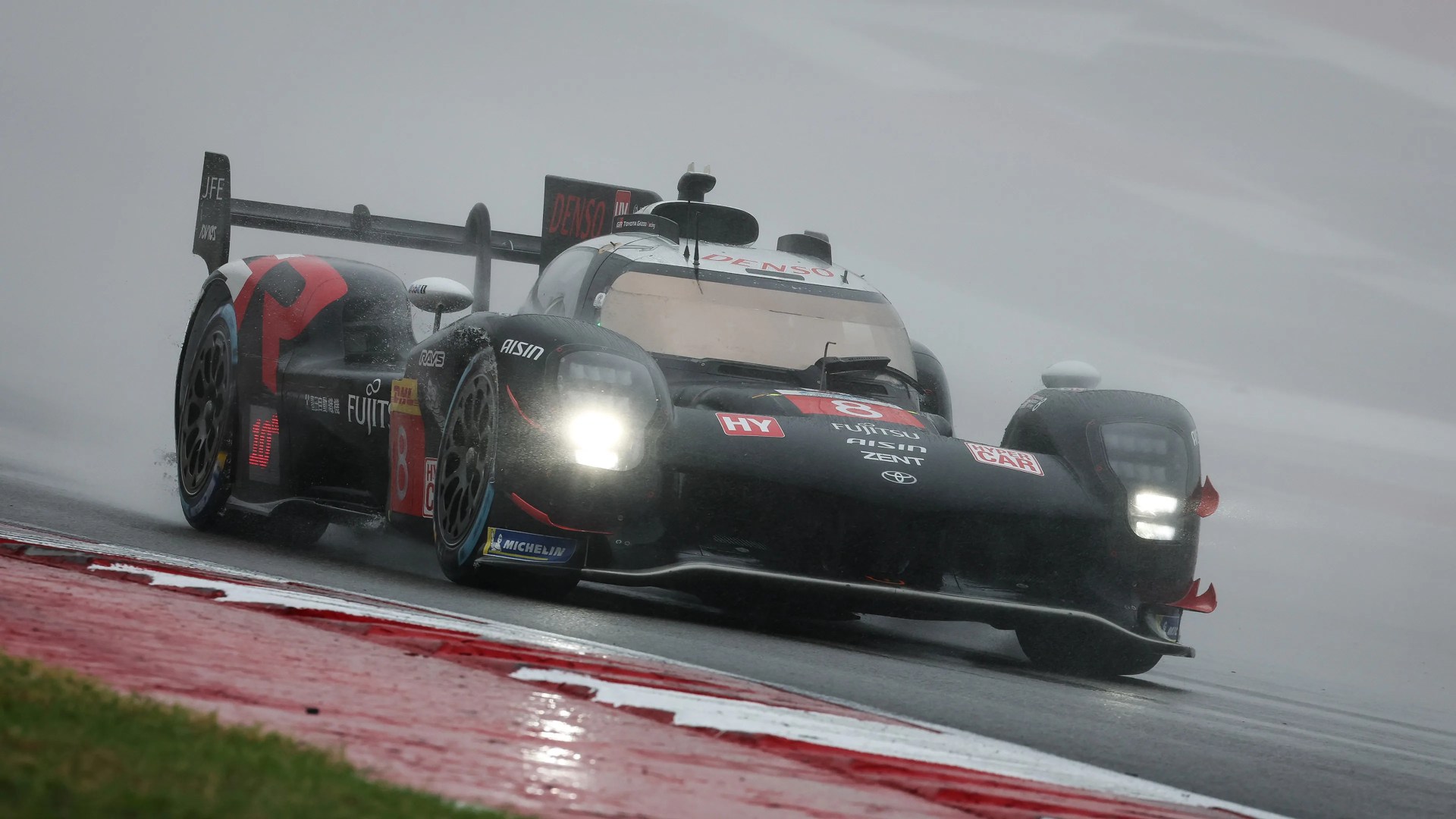 A black and red Toyota hybrid race car with the number 8 on the front, driving on a wet race track in rainy conditions with headlights on. The car features sponsor logos including Denso, Fujitsu, Aisin, and Michelin. The background is foggy and the track surface is wet and reflective.