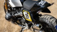 Close-up of the rear section of a black motorcycle with a gray seat, featuring the number "35" on a yellow-bordered white panel and the rear fender. The motorcycle has a visible exhaust pipe and a rugged tire.