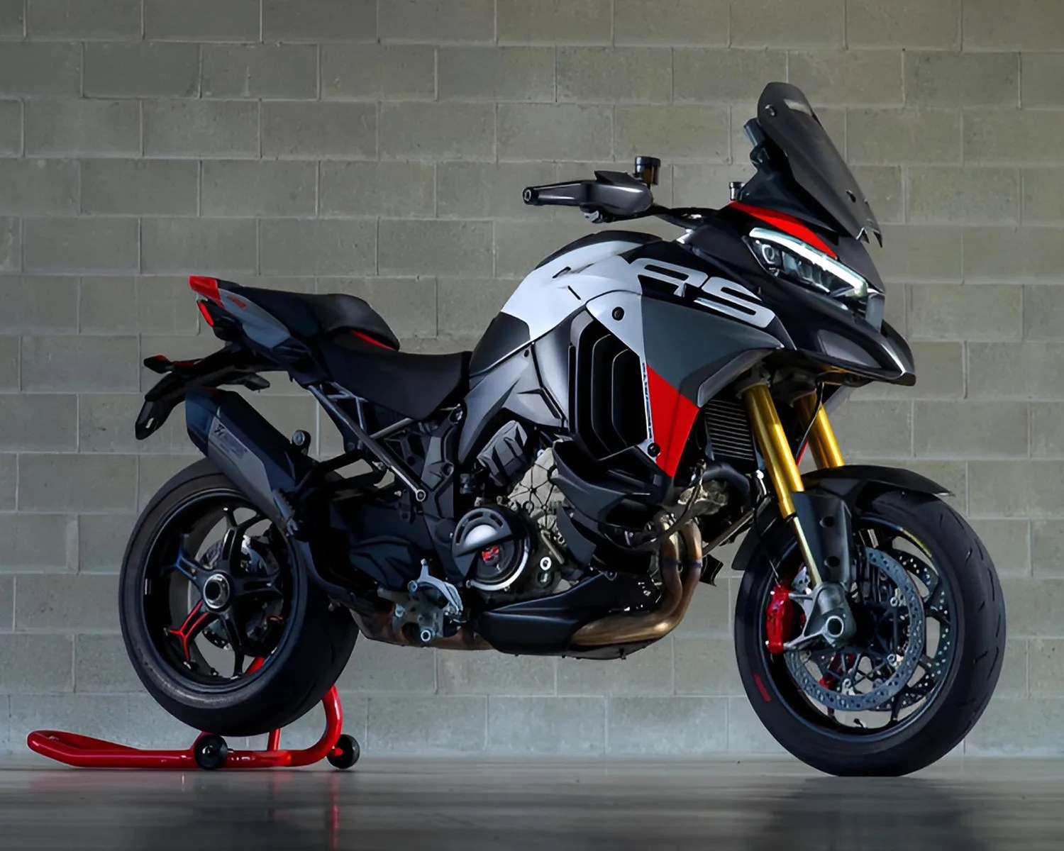 Sport motorcycle with a black, white, red, and gray color scheme, featuring "RS" branding on the side. It has gold front forks, a tinted windshield, and is supported by a red rear stand. The bike is positioned against a gray brick wall background.