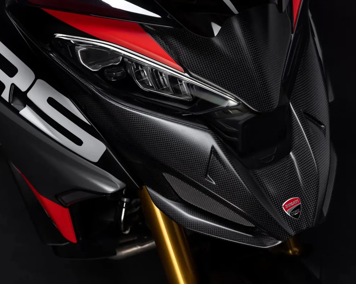 Close-up of a motorcycle front fairing featuring carbon fiber texture, a sleek headlight, red and black accents, and a Ducati Corse emblem. The front suspension forks are gold-colored.