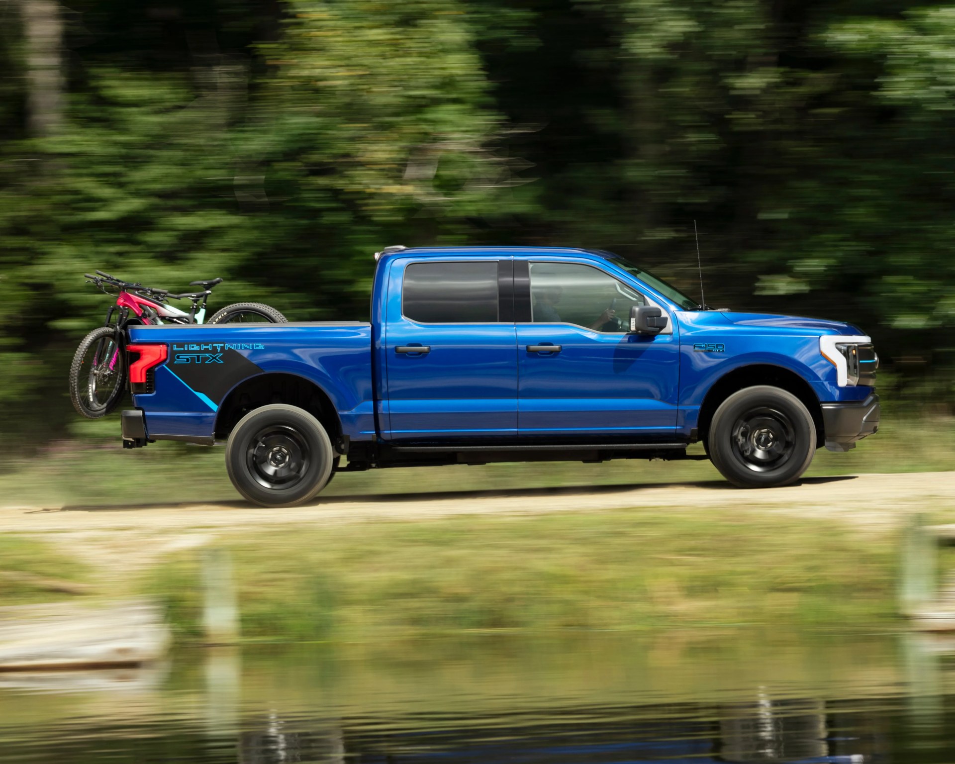 Blue Ford F-150 Lightning STX pickup truck driving on a dirt road with two mountain bikes mounted in the truck bed. The background is blurred greenery, indicating motion.