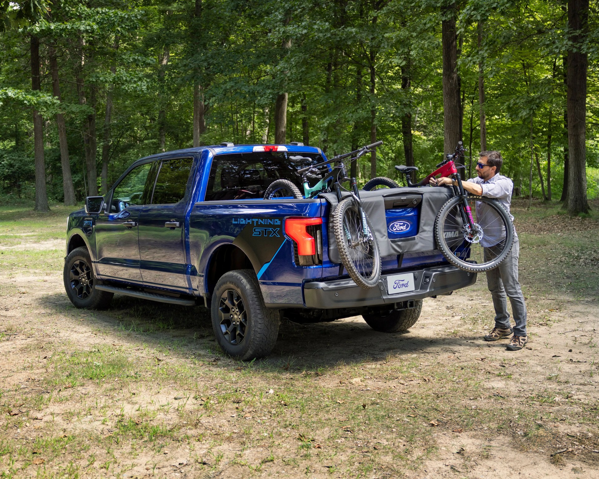 Blue Ford Lightning STX pickup truck parked in a wooded area with two mountain bikes mounted on the open tailgate. A man wearing sunglasses, a light-colored shirt, gray pants, and hiking shoes is adjusting one of the bikes. The tailgate has a protective pad with the Ford logo.