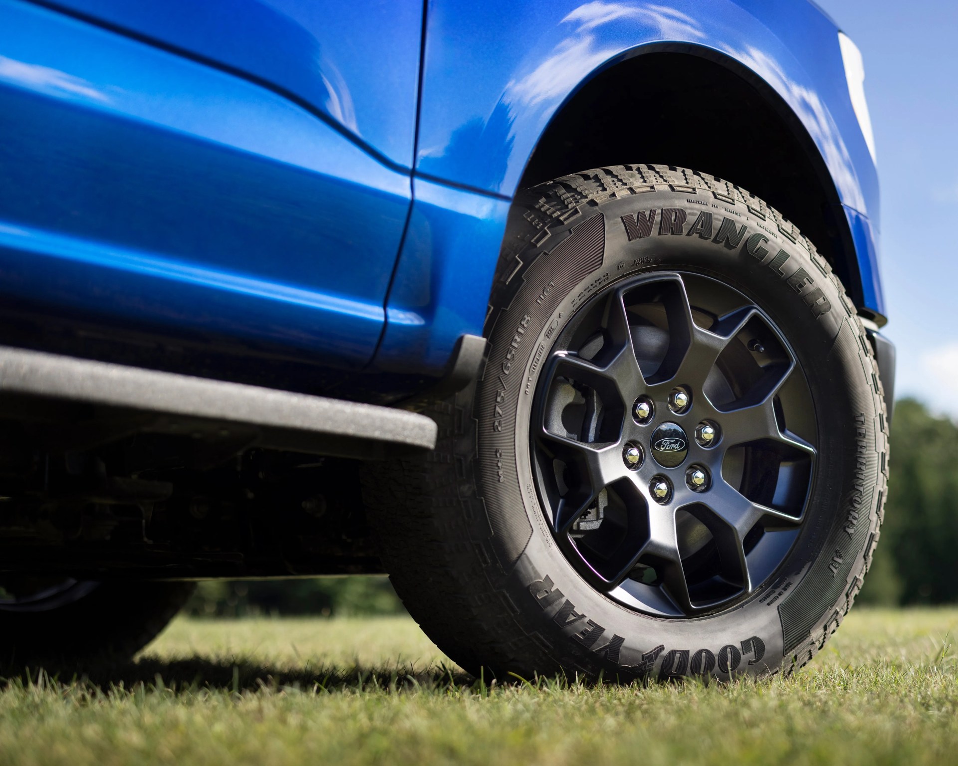 Close-up of a black Ford wheel with Goodyear Wrangler tire on a blue vehicle, parked on grass with a blurred natural background.