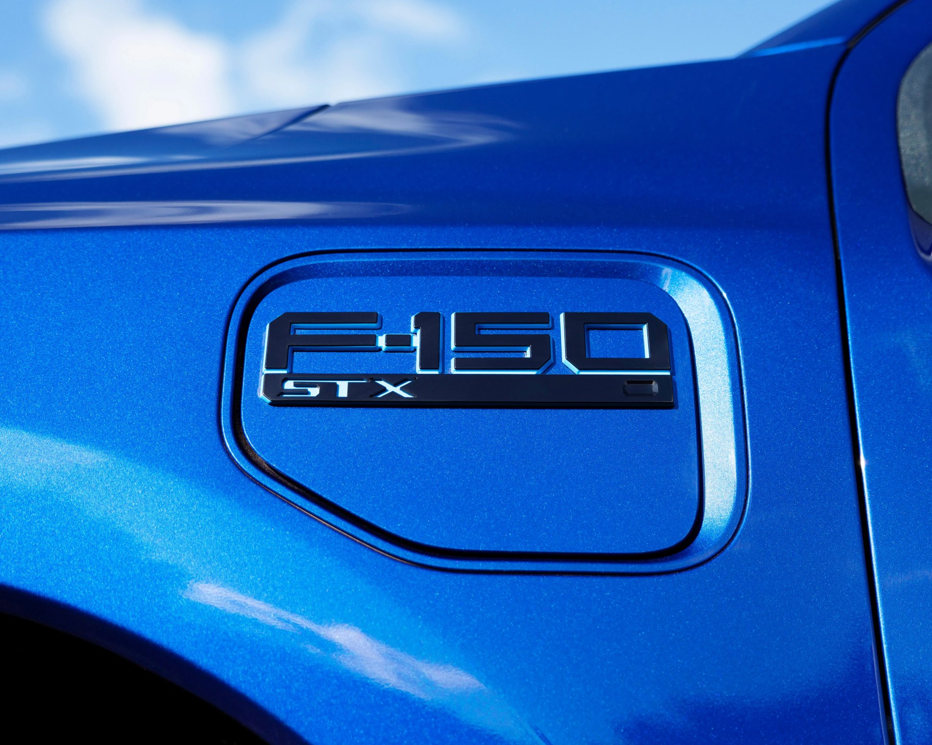 Close-up of a blue Ford F-150 truck's fuel door with a black "F-150 STX" emblem. The paint has a metallic finish, and the background shows a partly cloudy sky.