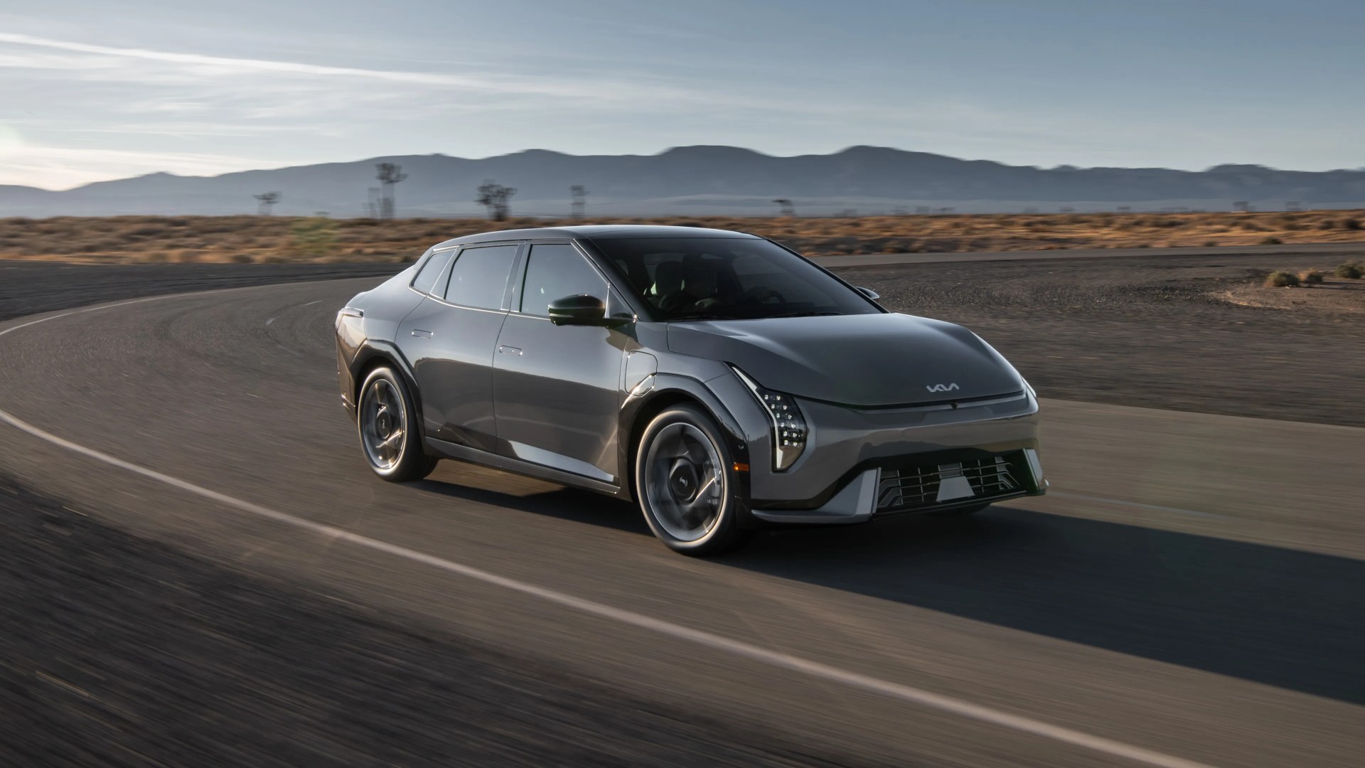 A sleek, modern gray electric car with a futuristic design is driving on a curved road in a desert landscape. The car features distinctive vertical LED headlights, a smooth aerodynamic body, and a black roof. Mountains and sparse desert vegetation are visible in the background under a clear sky.