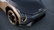 Front corner view of a modern Kia electric vehicle, showcasing a sleek gray hood with the Kia logo, angular LED headlights, and a black and silver alloy wheel with Nexen tires, parked on an asphalt surface.
