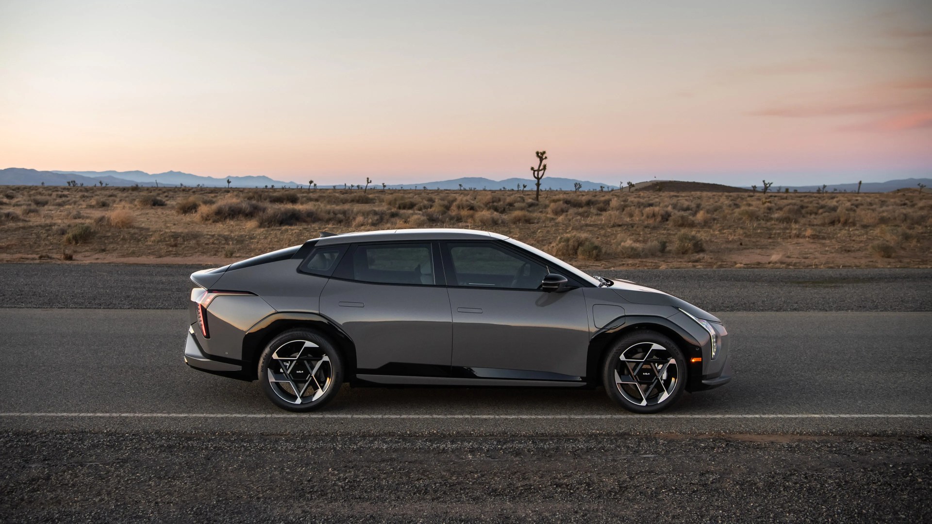 A sleek, modern gray electric SUV with black accents is parked on a desert road at sunset. The vehicle features sharp, angular design elements, distinctive geometric alloy wheels, and a streamlined profile. The background shows a dry, arid landscape with sparse vegetation and distant mountains under a soft, pastel-colored sky.
