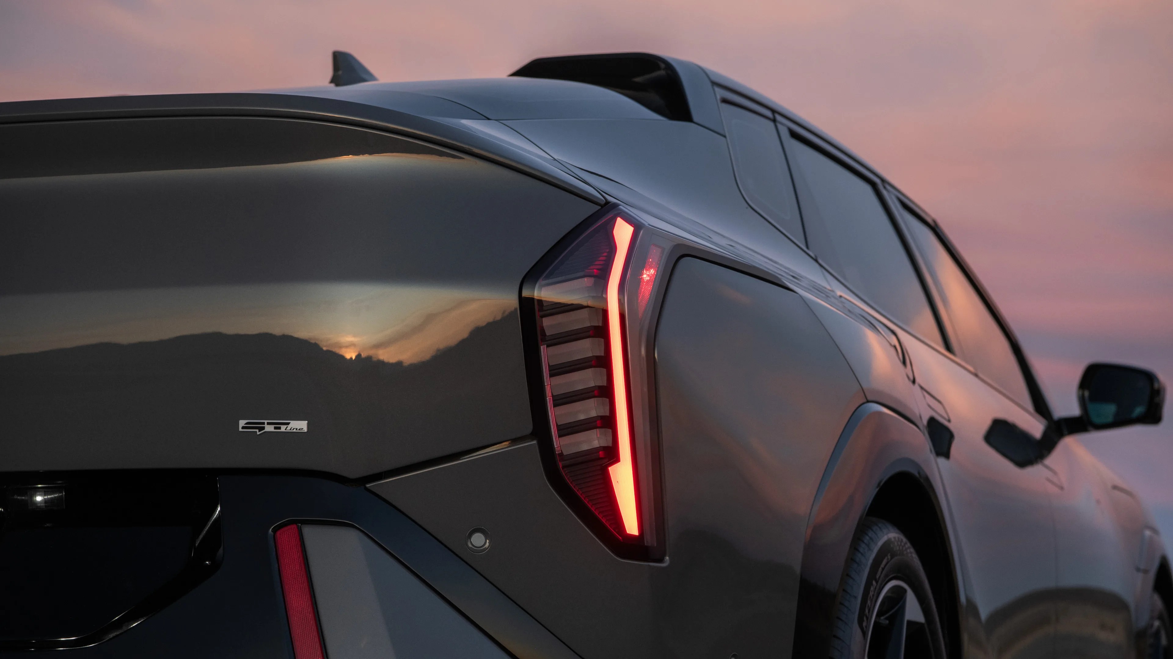 Rear side view of a dark-colored modern SUV at sunset, highlighting the illuminated vertical tail light and the "GT Line" badge on the tailgate. The vehicle's glossy surface reflects the sunset and surrounding landscape.