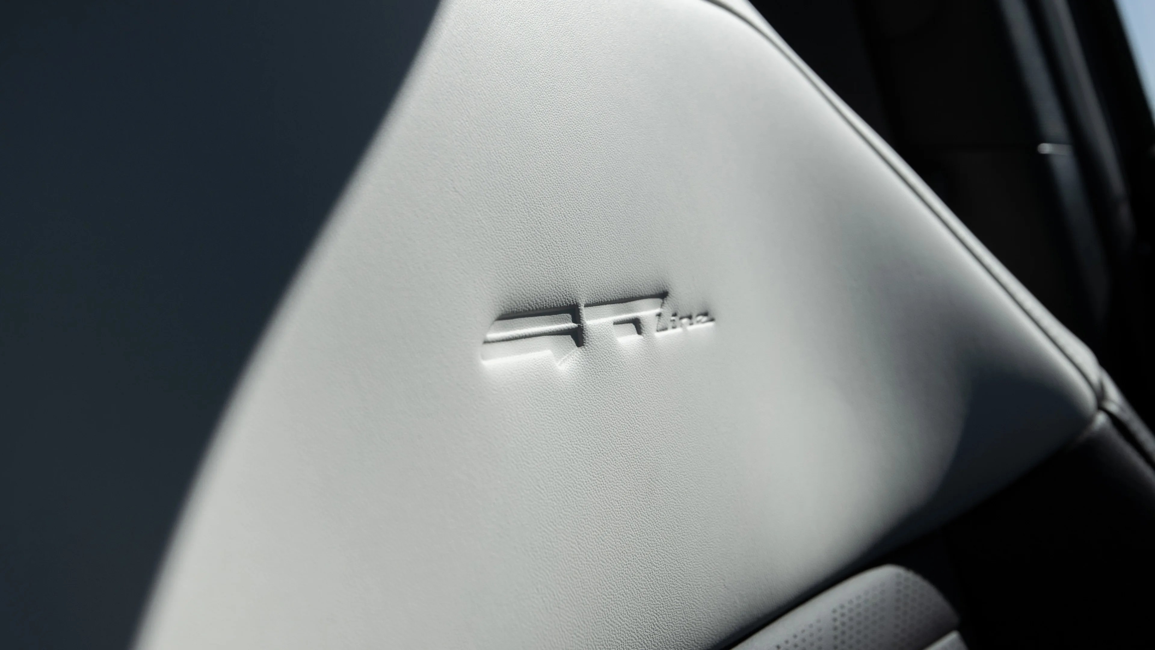 Close-up of a light gray leather car seat with the embossed "GT line" logo in the center. The leather has a smooth texture with visible stitching along the edge. Shadows partially cover the seat, adding depth to the image.