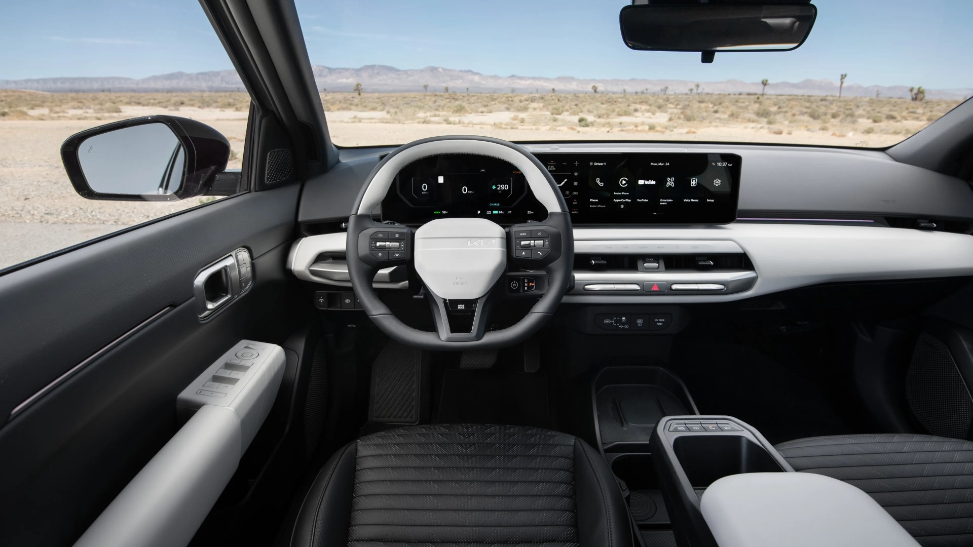 Modern car interior featuring a black and white steering wheel with integrated controls, a digital instrument cluster, and a wide touchscreen infotainment display. The dashboard has a sleek, minimalist design with horizontal air vents and a combination of black and white surfaces. The driver's door panel includes window and mirror controls, and the seats are black with a textured pattern. The view outside shows a desert landscape with mountains in the distance.