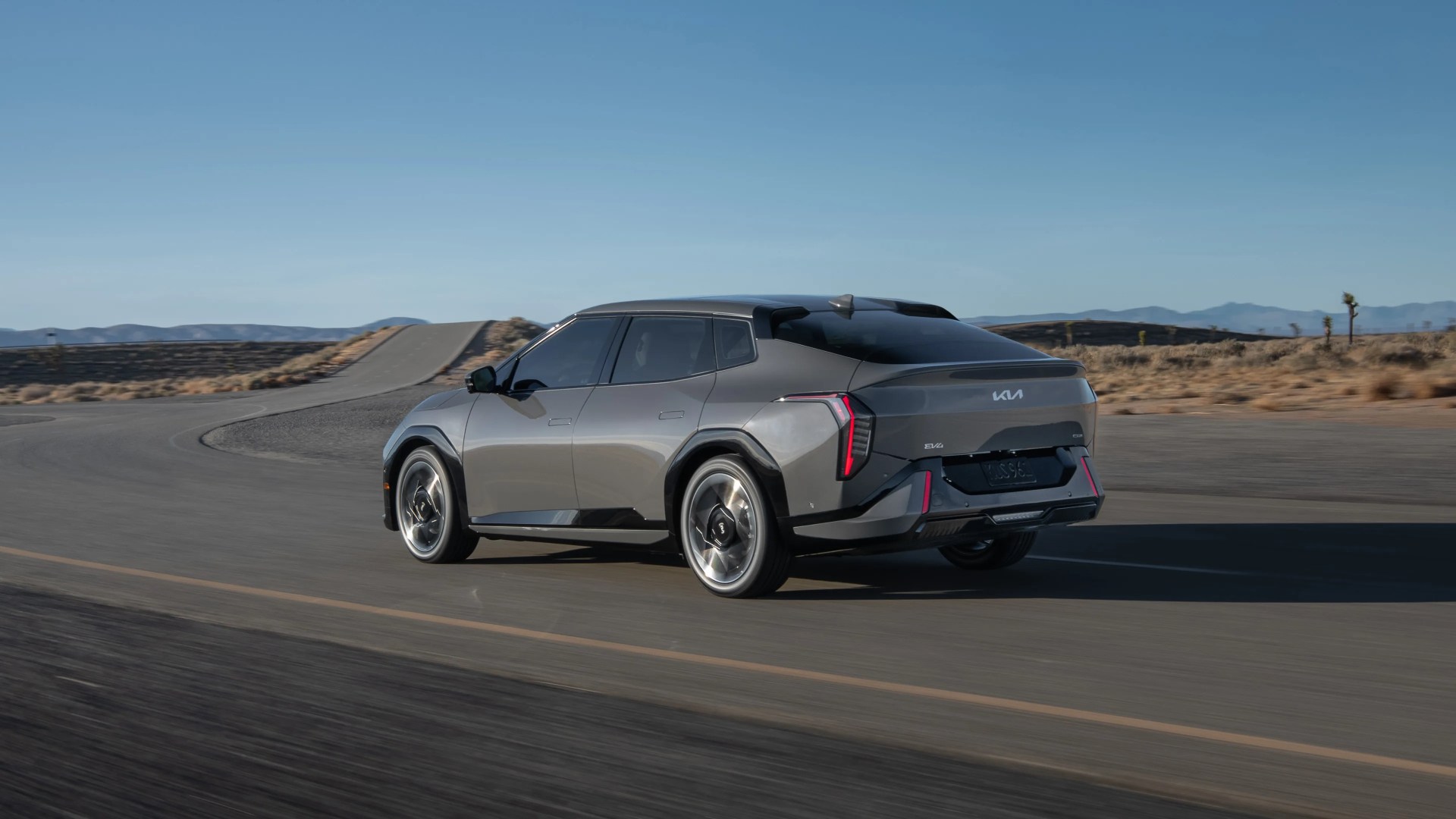 Gray Kia EV6 electric crossover driving on a paved road in a desert landscape with clear blue sky. The car has a sleek, modern design with distinctive rear lights and black accents.