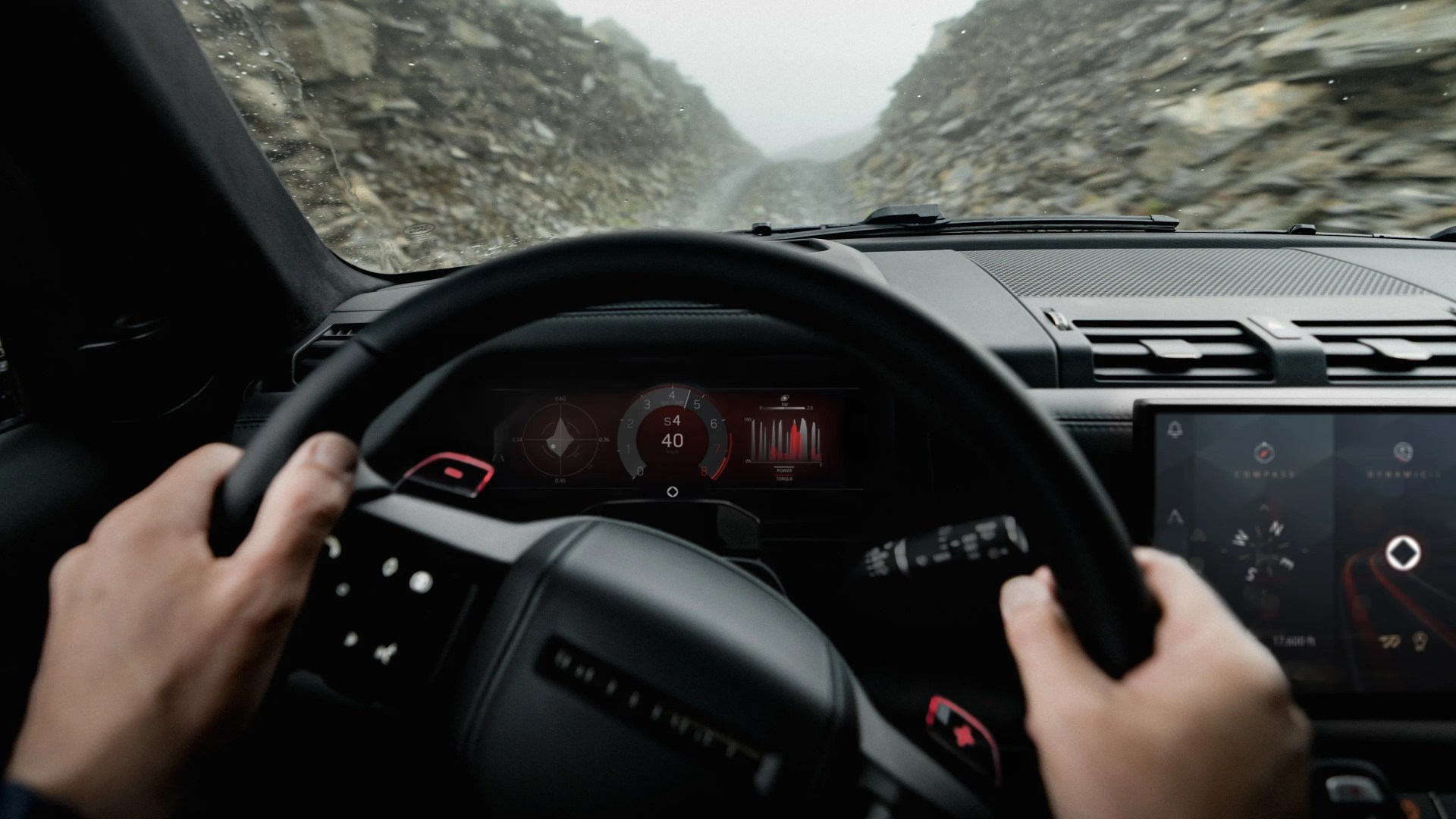 View from inside a vehicle showing hands on a steering wheel, a digital dashboard displaying speed and other metrics, and a touchscreen navigation system. Outside the windshield, a narrow, rocky dirt road is visible between steep rock walls, with a misty or foggy atmosphere.