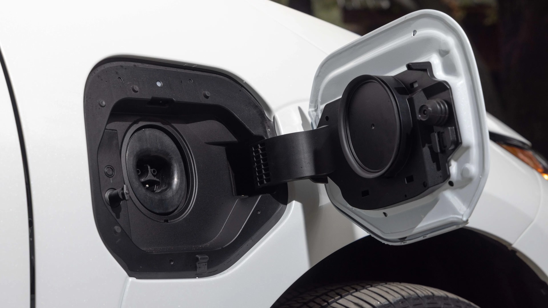 Open fuel filler door on a white vehicle, showing the black fuel cap area and the hinge connecting the door to the car body. The tire and part of the wheel well are visible below.