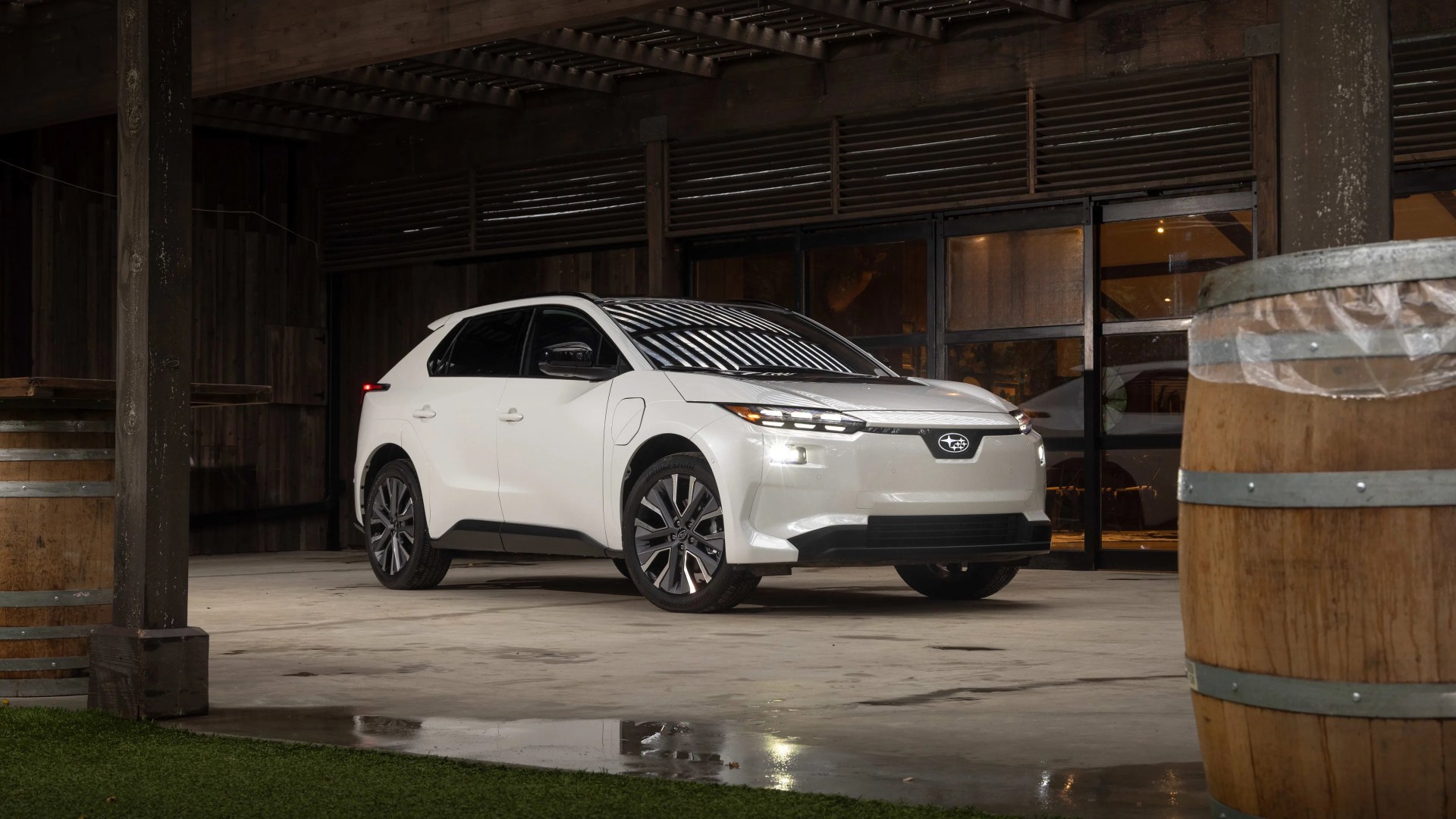 White Subaru electric SUV parked indoors on a concrete floor with wooden barrels and a wooden structure in the background. The vehicle has a sleek, modern design with black accents and illuminated headlights.