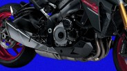 Close-up of the engine and exhaust system of a Suzuki GSX-S 1000 motorcycle, featuring a black frame, red wheels, and gold front forks. The Brembo brake caliper is visible on the front wheel.