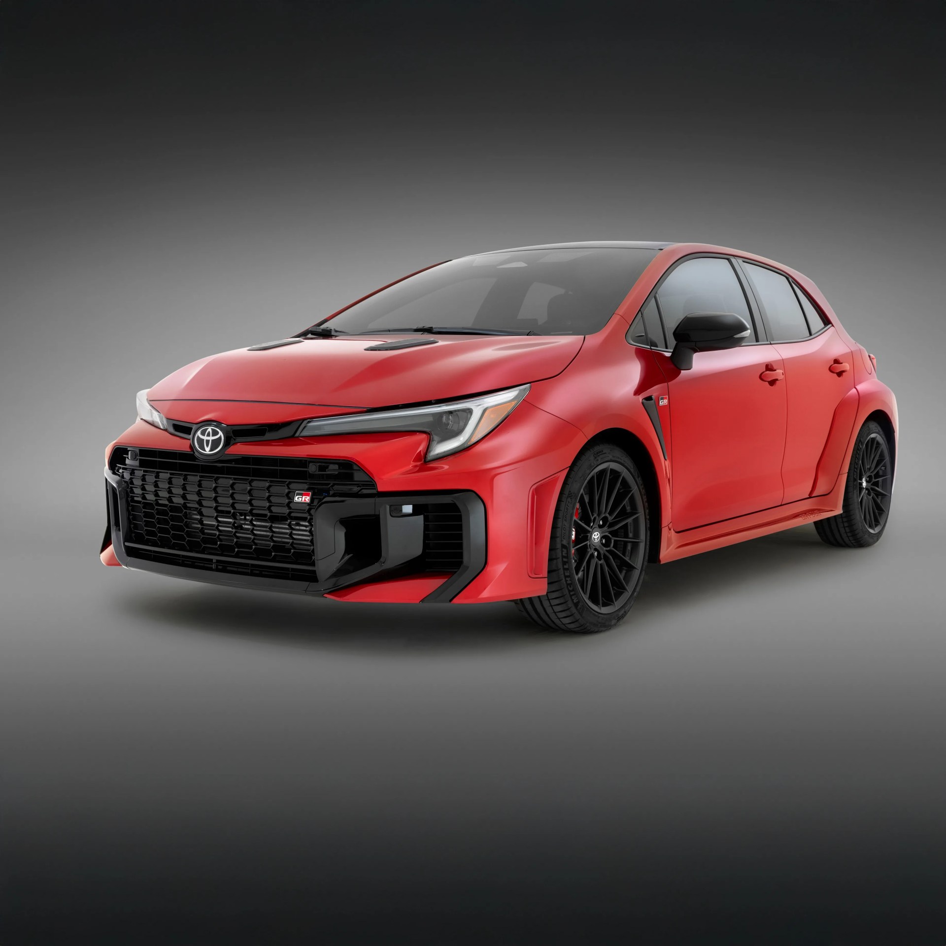 Red Toyota GR hatchback with black accents, including a large black front grille, black side mirrors, and black multi-spoke wheels, shown at a three-quarter front angle on a gray gradient background.