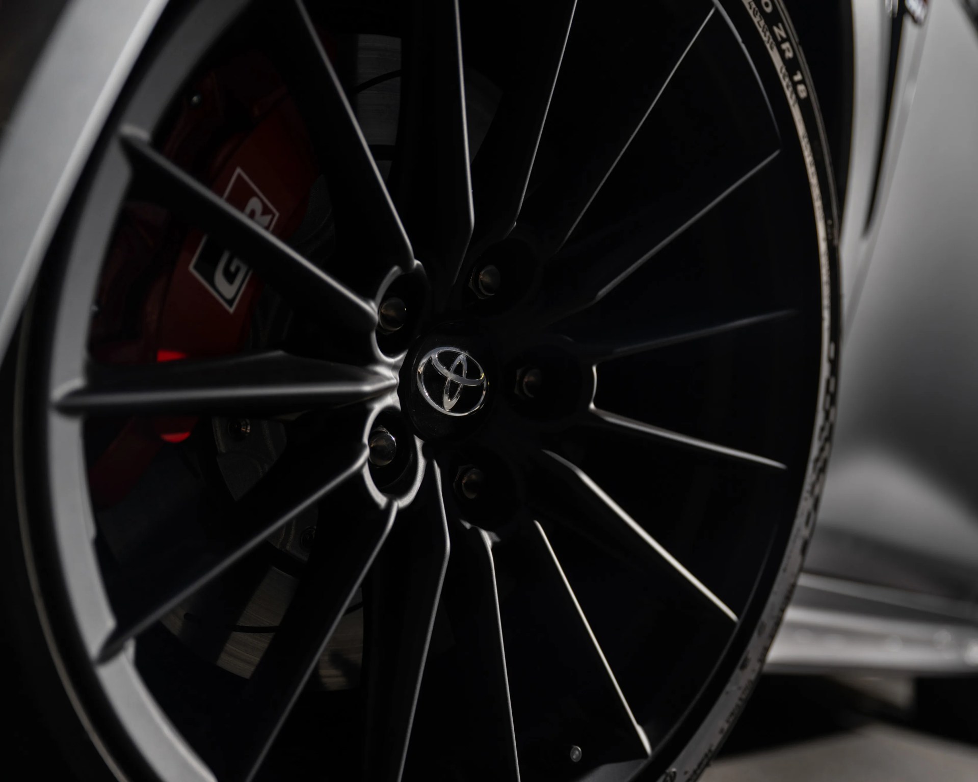 Black multi-spoke alloy wheel with a Toyota logo in the center, featuring a red brake caliper with "GR" branding visible behind the spokes.
