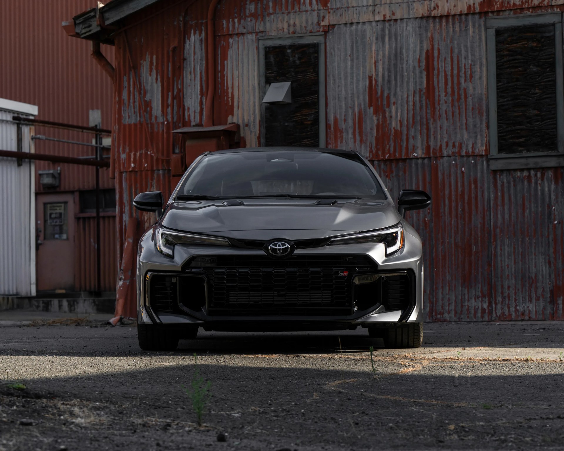 Front view of a silver Toyota GR car parked on a rough asphalt surface in front of a weathered, rusted corrugated metal building. The car features sleek, angular headlights and a prominent black grille with the Toyota emblem and GR badge.