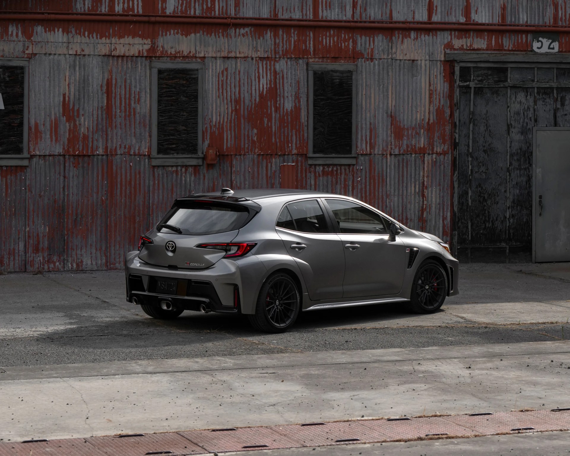 A silver Toyota GR Corolla hatchback is parked in front of a weathered, corrugated metal building with peeling red and gray paint. The car features black wheels, tinted windows, and sporty design elements including a rear spoiler and dual exhaust pipes. The setting appears industrial and slightly rundown.