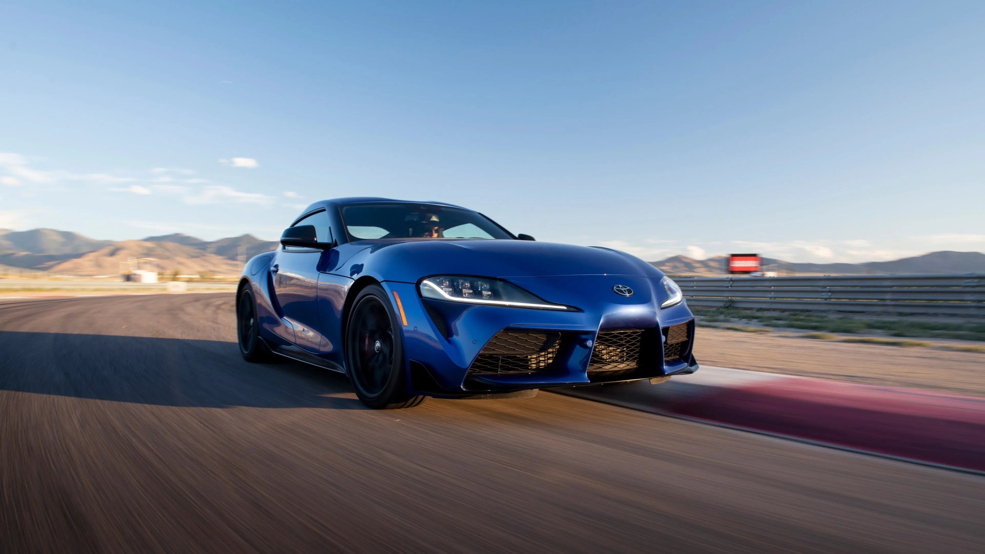 Blue Toyota Supra sports car driving on a racetrack with mountains in the background under a clear sky.