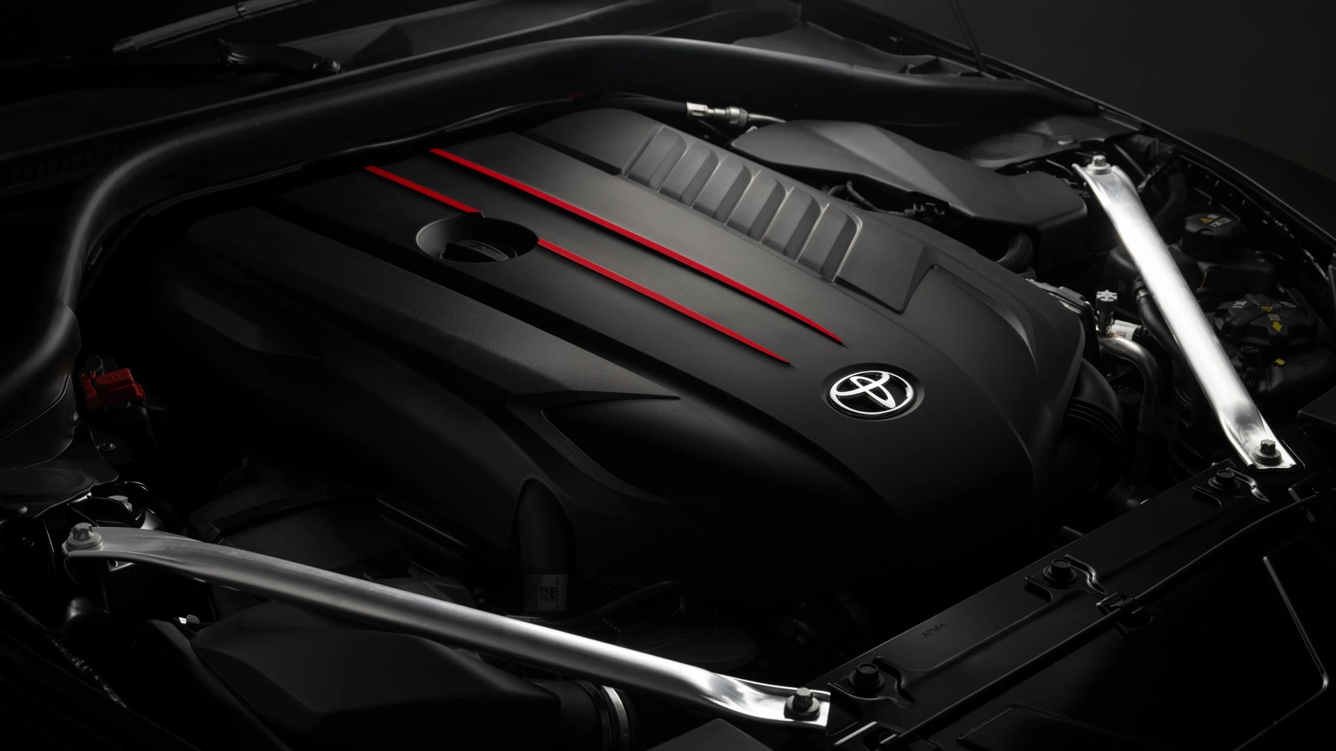 Close-up of a Toyota car engine with a sleek black cover featuring three red accent lines and the Toyota emblem in the center, surrounded by metallic support bars and various engine components.
