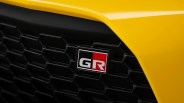 Close-up of a black honeycomb car grille with a "GR" emblem in the center, featuring a black and red background, set against a yellow car body.