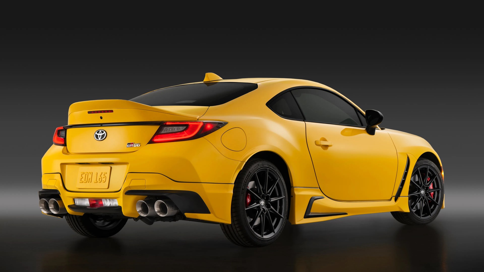 Yellow Toyota GR86 sports car viewed from the rear three-quarter angle, featuring black alloy wheels, quad exhaust tips, a rear spoiler, and tinted windows against a dark gradient background.