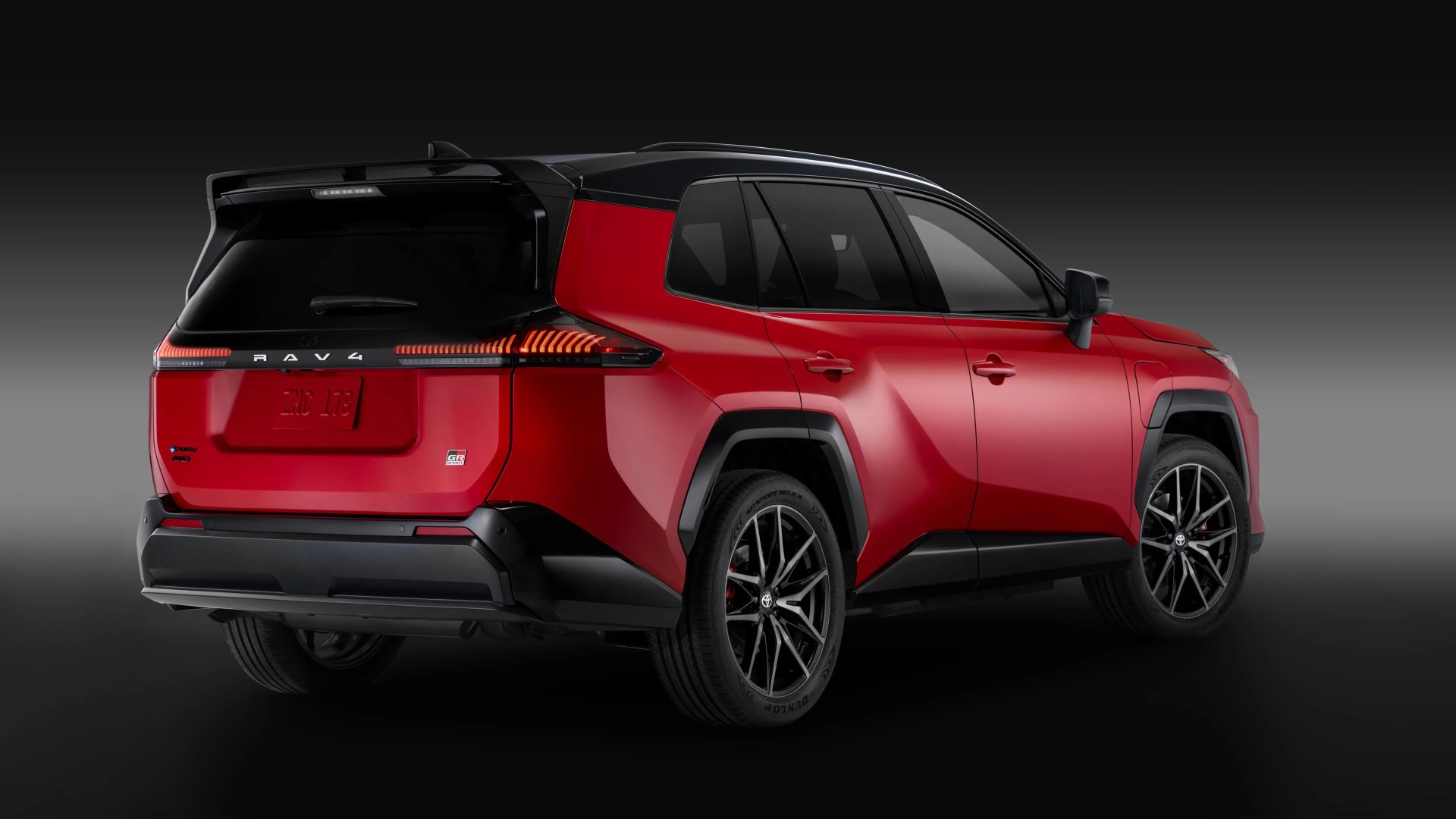 Red Toyota RAV4 GR Sport SUV with black roof and black trim, shown from the rear three-quarter angle against a gradient dark background. The vehicle features sleek, modern taillights and sporty black alloy wheels.
