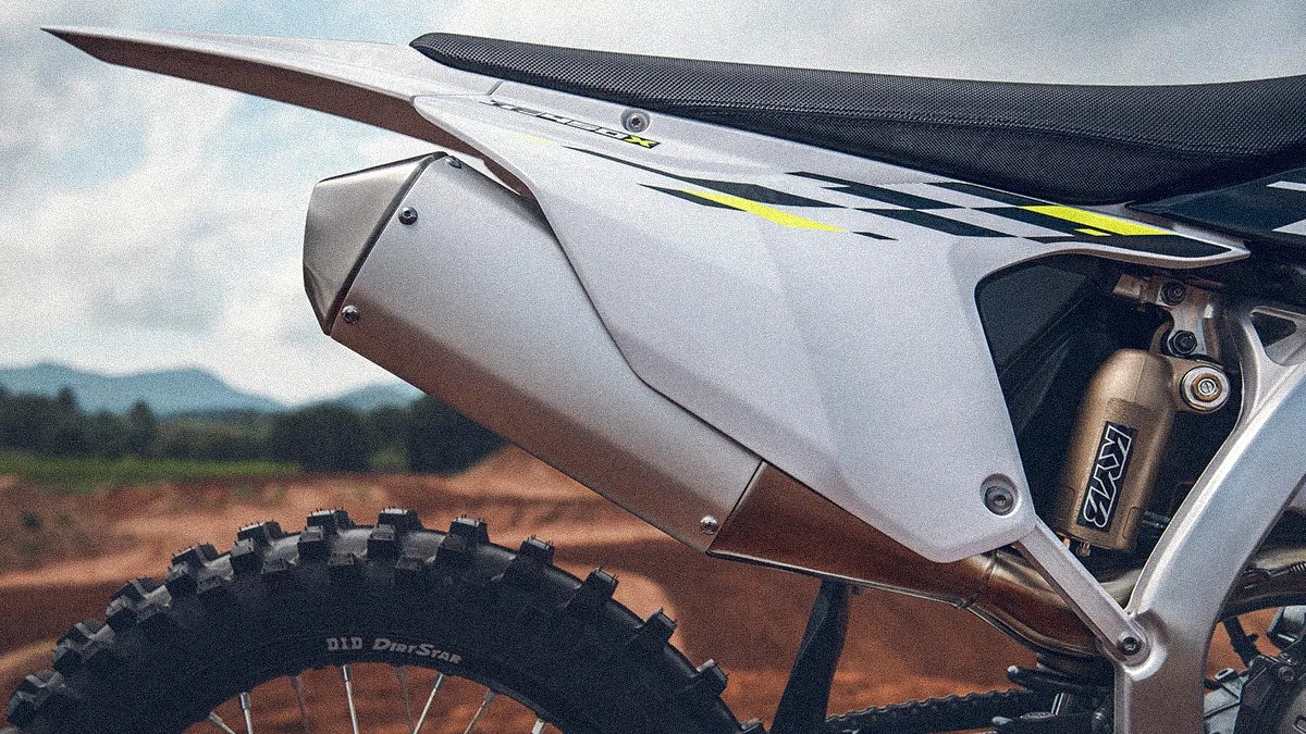 Close-up of the rear section of a dirt bike, showing the exhaust pipe, rear suspension with a KYB shock absorber, part of the seat, and the knobby rear tire. The bike has a white and black color scheme with yellow accents. The background features an outdoor dirt track with blurred natural scenery.
