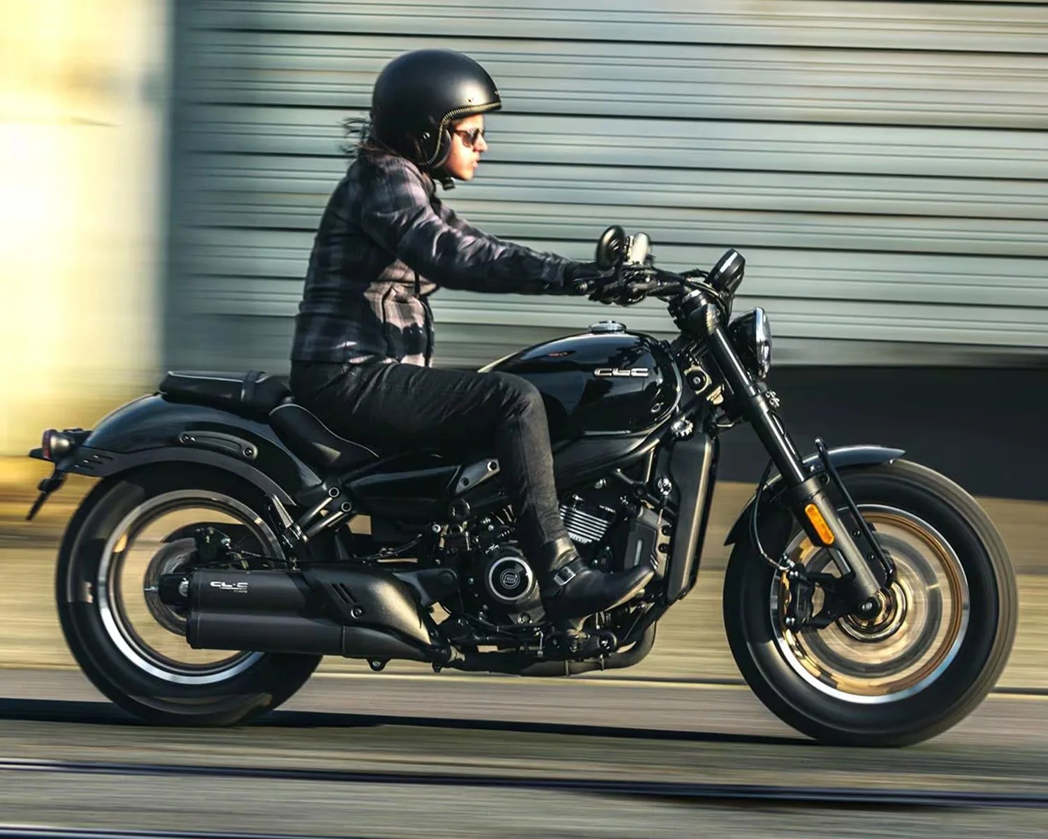 A person wearing a black helmet, sunglasses, a dark plaid jacket, black pants, and black boots is riding a black motorcycle with the letters "CLC" on the fuel tank and exhaust. The motorcycle is in motion, with the wheels blurred to indicate speed. The background features a horizontal metal shutter.