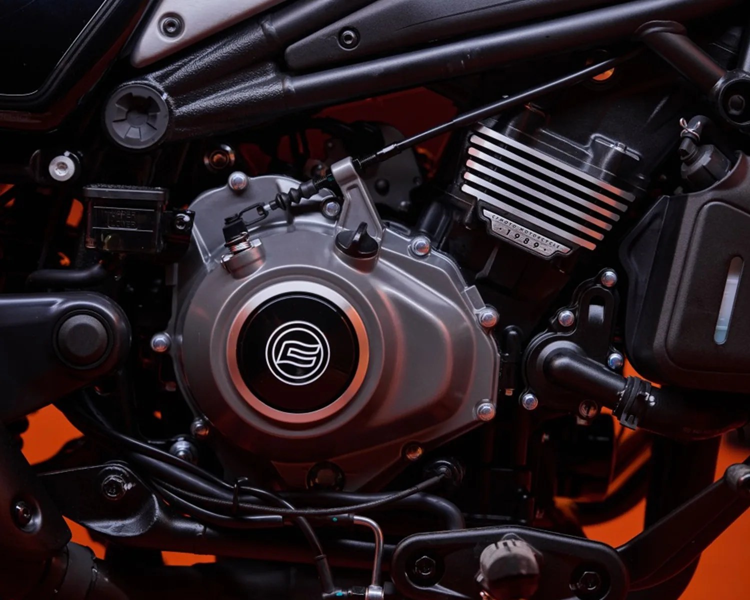 Close-up of a motorcycle engine showing the metallic engine casing with a circular emblem in the center, black and silver components, bolts, and a label reading "CFMOTO MOTORCYCLE 1989" on the finned cylinder head. The background has a warm orange glow.