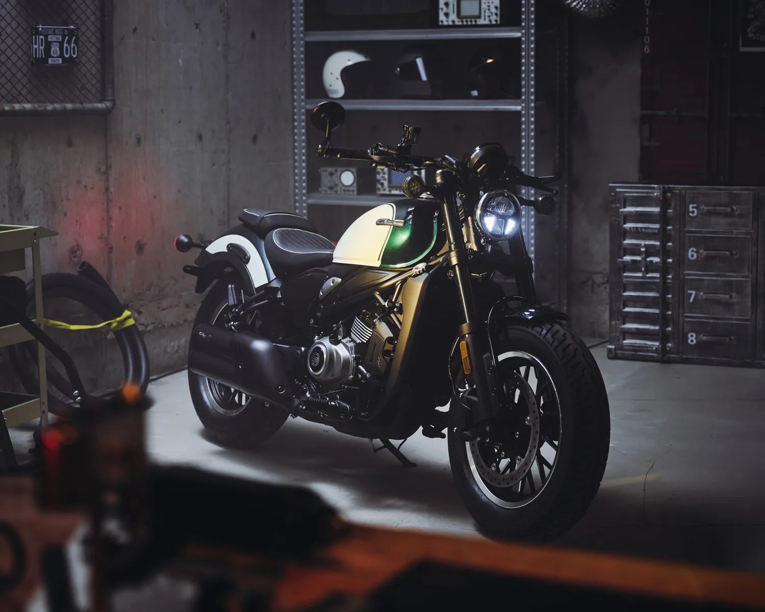 A black and white motorcycle with a green fuel tank is parked indoors on a concrete floor. The motorcycle has a round headlight turned on, black exhaust, and a black seat with a textured pattern. In the background, there is a metal shelving unit with helmets and other items, a concrete wall, and a metal cabinet with numbered drawers. A tire is leaning against the wall on the left side.