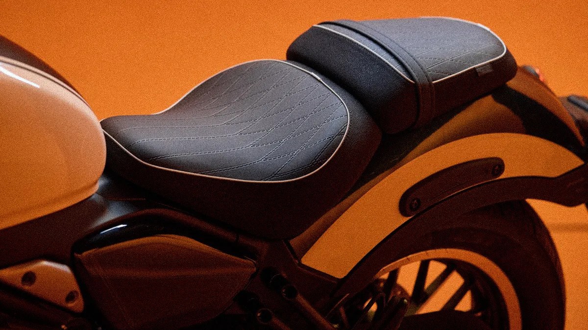 Black leather motorcycle seat with detailed stitching, featuring a larger front seat and a smaller rear seat, set against an orange background.