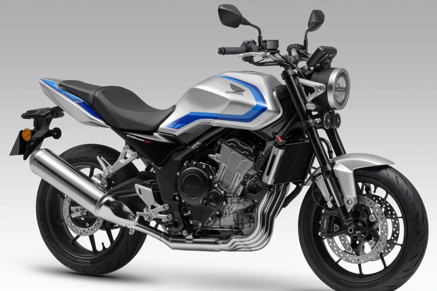 Silver and black Honda motorcycle with blue accents, featuring a round headlight, dual front disc brakes, and a large exhaust pipe on the left side. The bike has a sporty design with a black seat and visible engine components.
