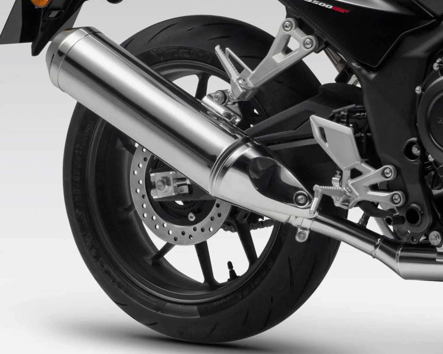 Rear wheel and exhaust system of a black motorcycle, featuring a large, shiny chrome exhaust pipe, a disc brake, and a black tire with visible tread. The motorcycle frame and footrest are also visible in silver and black.