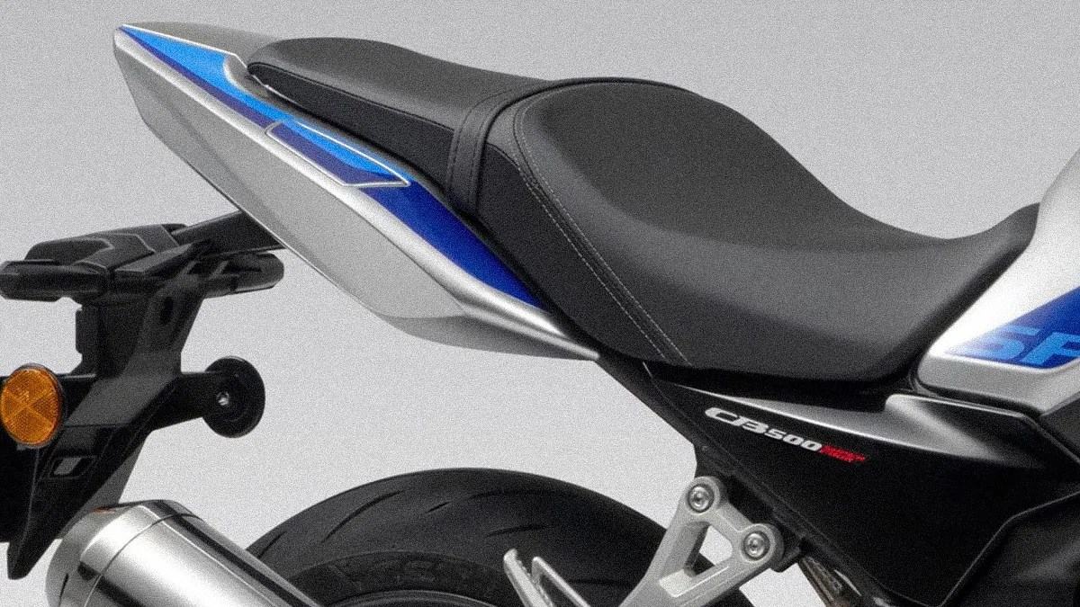 Rear section of a motorcycle featuring a black seat with detailed stitching, a blue and silver tail fairing, a rear tire, and an exhaust pipe. The model designation "CB500F" is visible on the black panel below the seat.