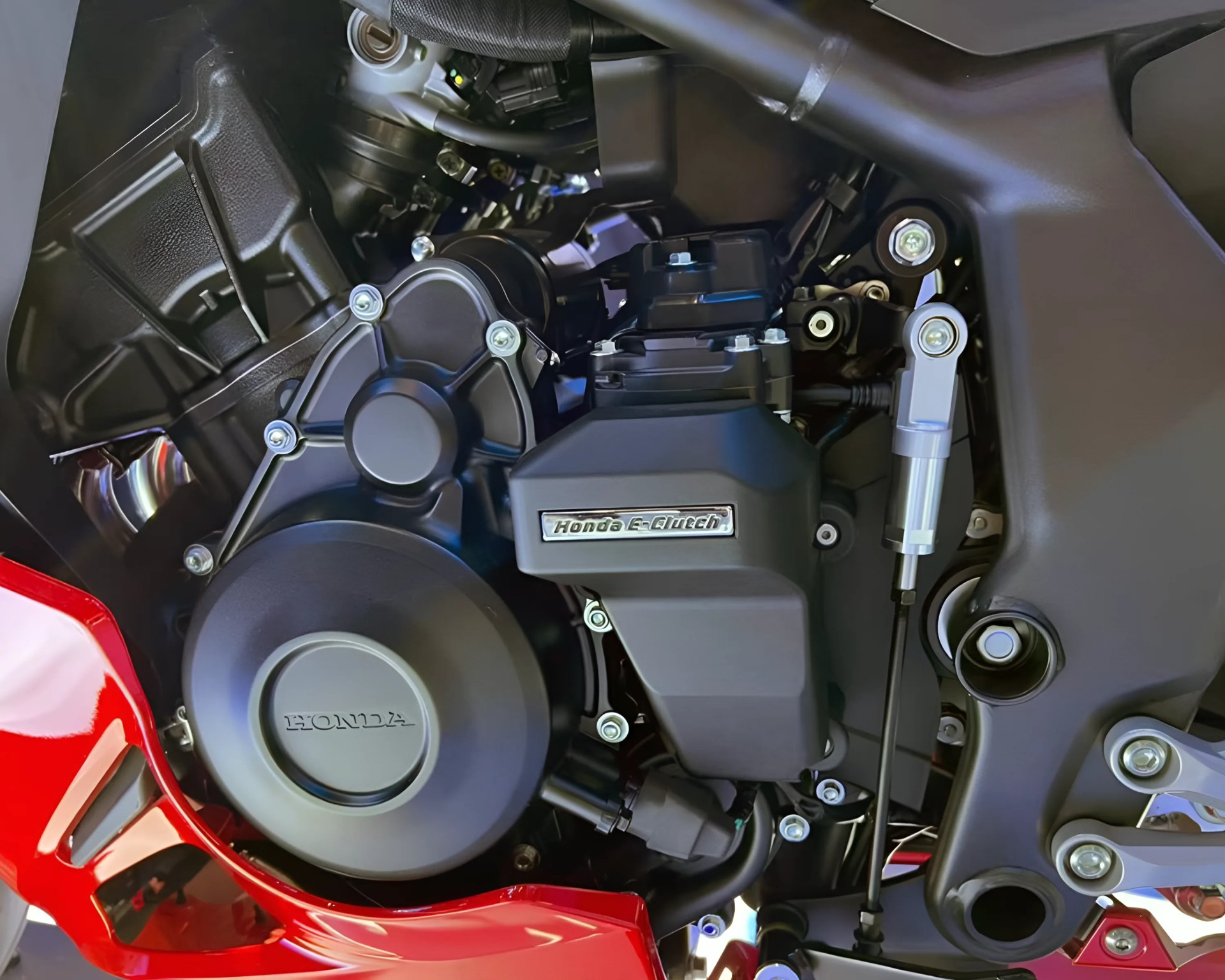 Close-up of a Honda motorcycle engine featuring the "Honda e:Architecture" label on a black casing, with visible bolts, mechanical components, and part of the red motorcycle frame.