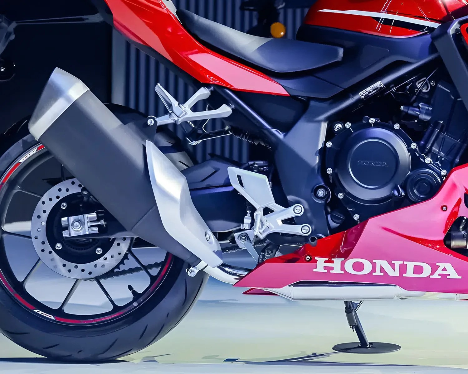 Close-up of the rear section of a red Honda motorcycle, showing the rear wheel with a disc brake, exhaust pipe, foot pegs, and part of the engine casing. The Honda logo is visible on the lower fairing.