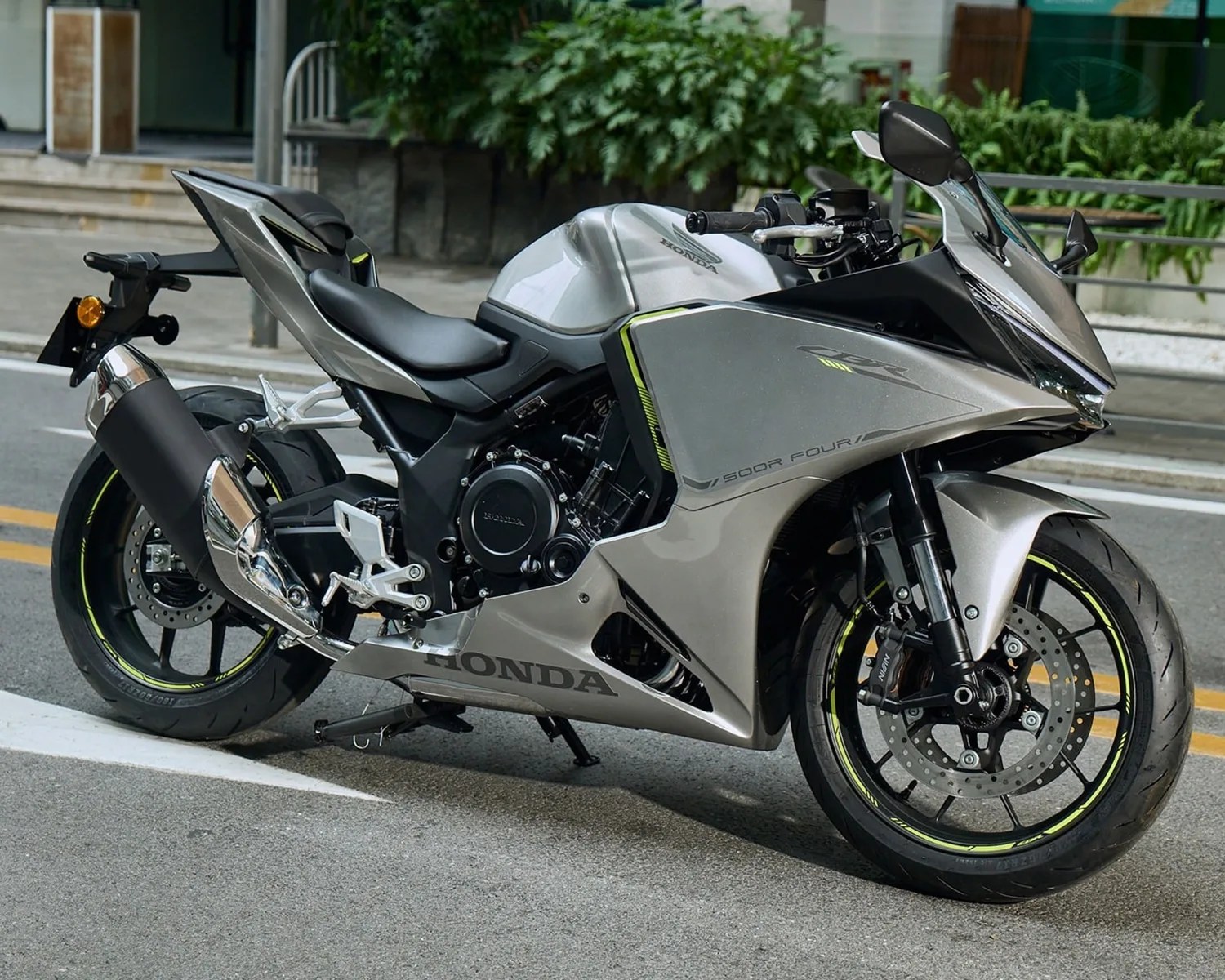 Silver Honda CBR 500R sport motorcycle parked on a street, featuring a sleek aerodynamic design with black and neon yellow accents on the wheels and fairings. The bike has a black seat, large exhaust pipe, and visible front disc brakes.