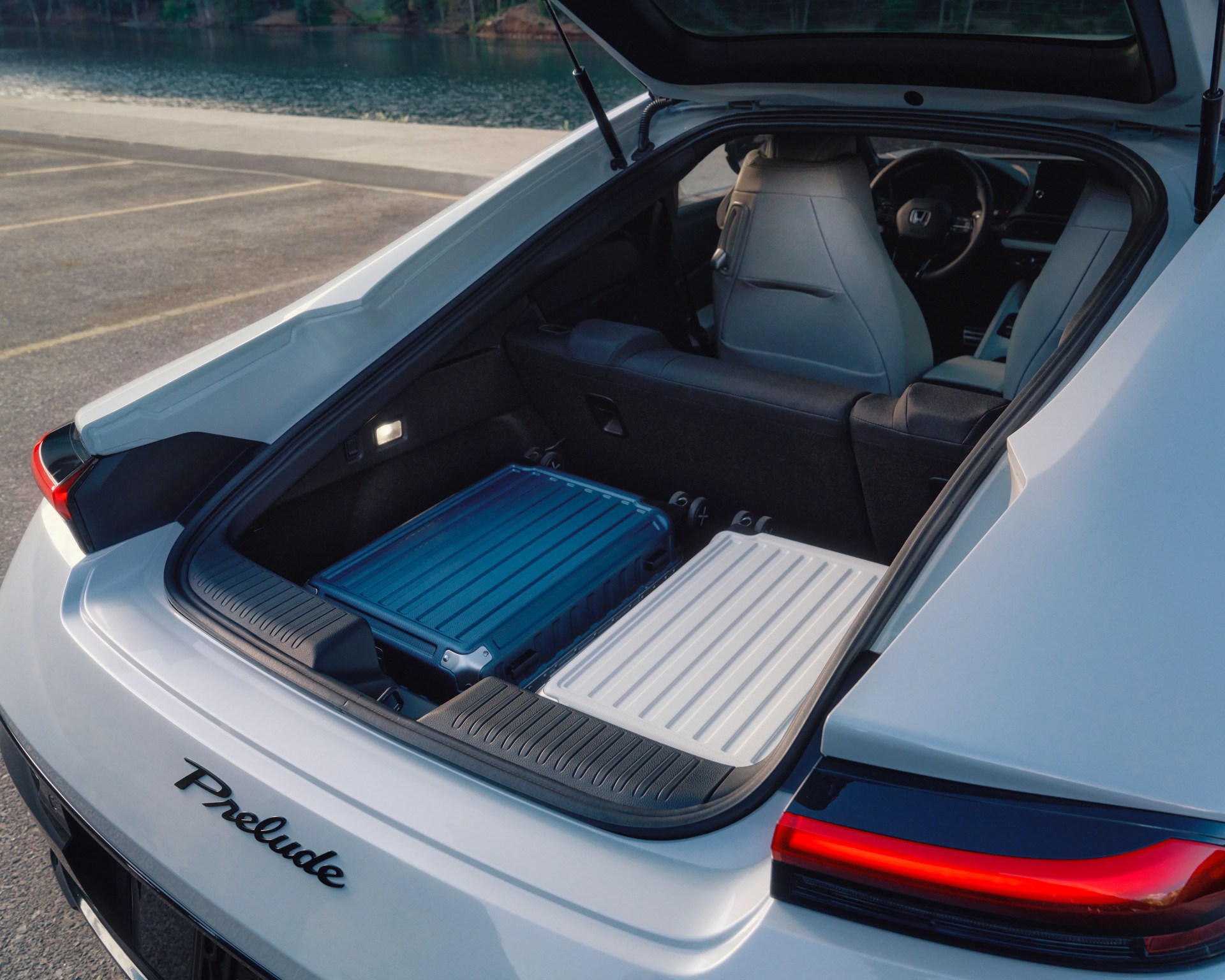 Open trunk of a white Honda Prelude showing two hard-shell suitcases, one blue and one white, inside. The interior features light-colored front seats and a visible steering wheel. The car is parked near a body of water.