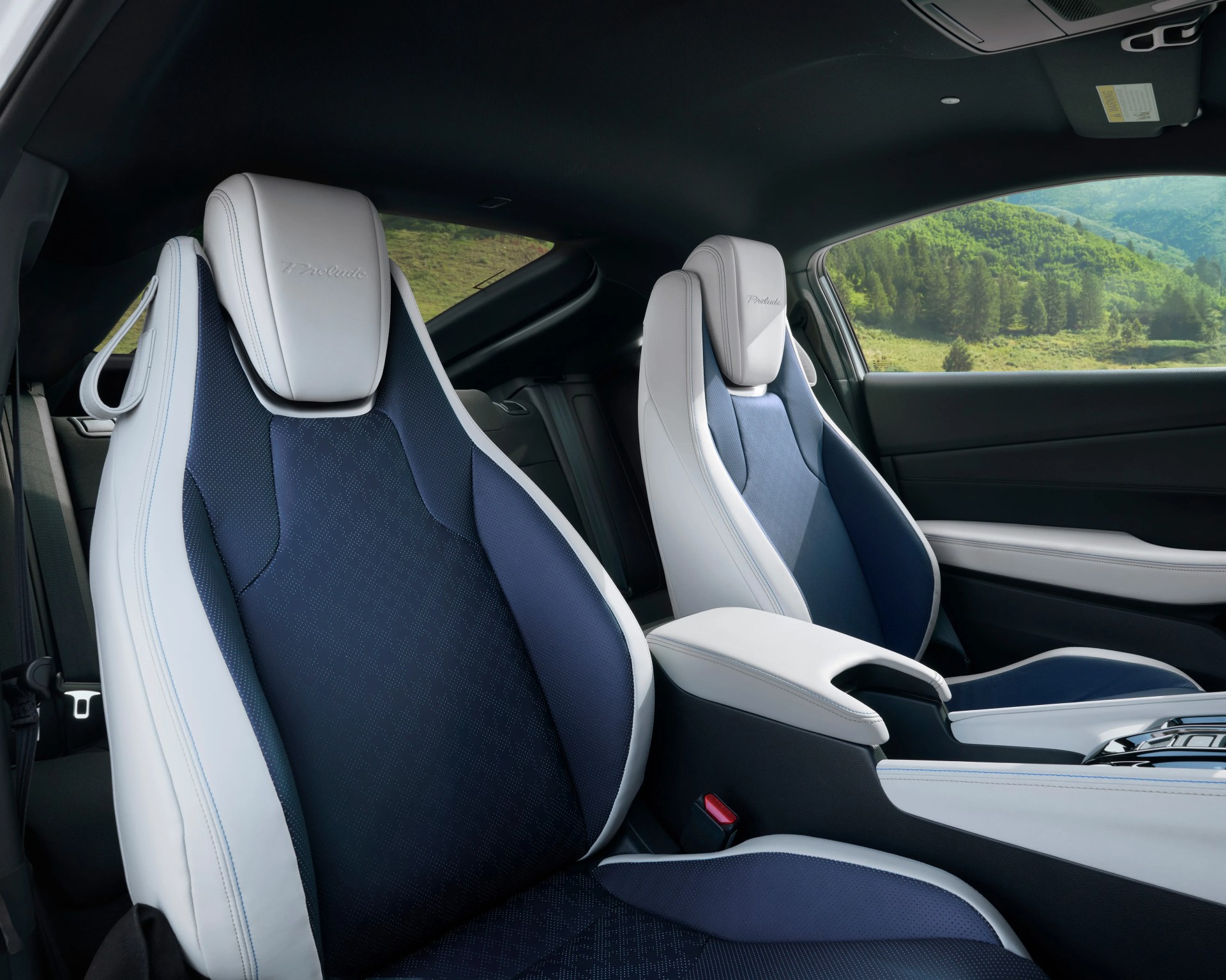 Two-tone car seats with white leather sides and navy blue fabric centers, featuring "Prelude" embroidered on the headrests. The interior includes a white and black center console, with a view of green hills and trees visible through the window.