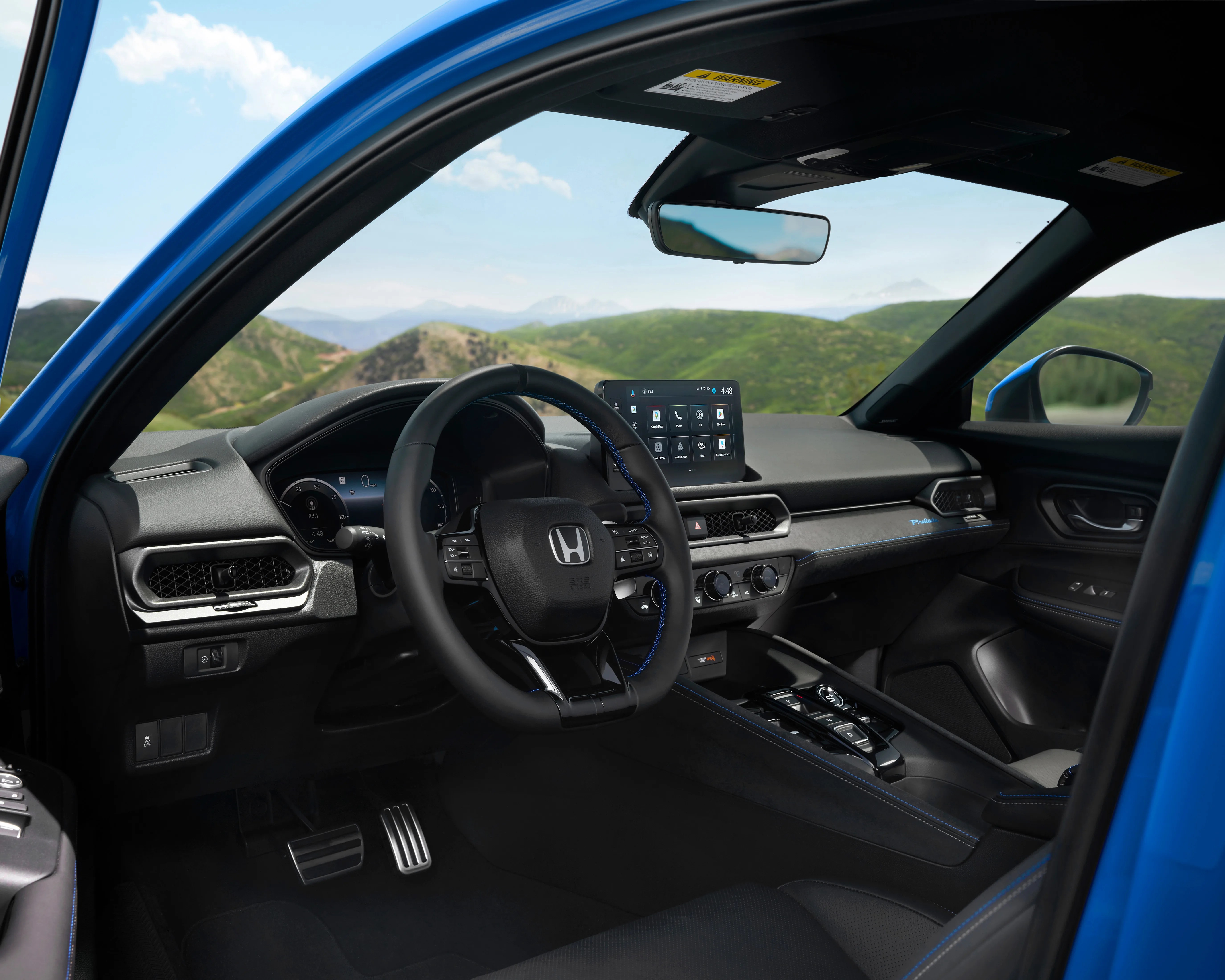 Interior of a modern Honda vehicle featuring a black steering wheel with blue stitching, a digital instrument cluster, and a central touchscreen display on the dashboard. The dashboard has a sleek design with black and dark gray materials, blue accent stitching, and a "Prologue" badge. The center console includes push-button gear selectors and climate control knobs. The driver's side door panel has window and mirror controls, and the view outside shows a scenic landscape with green hills and a partly cloudy sky.