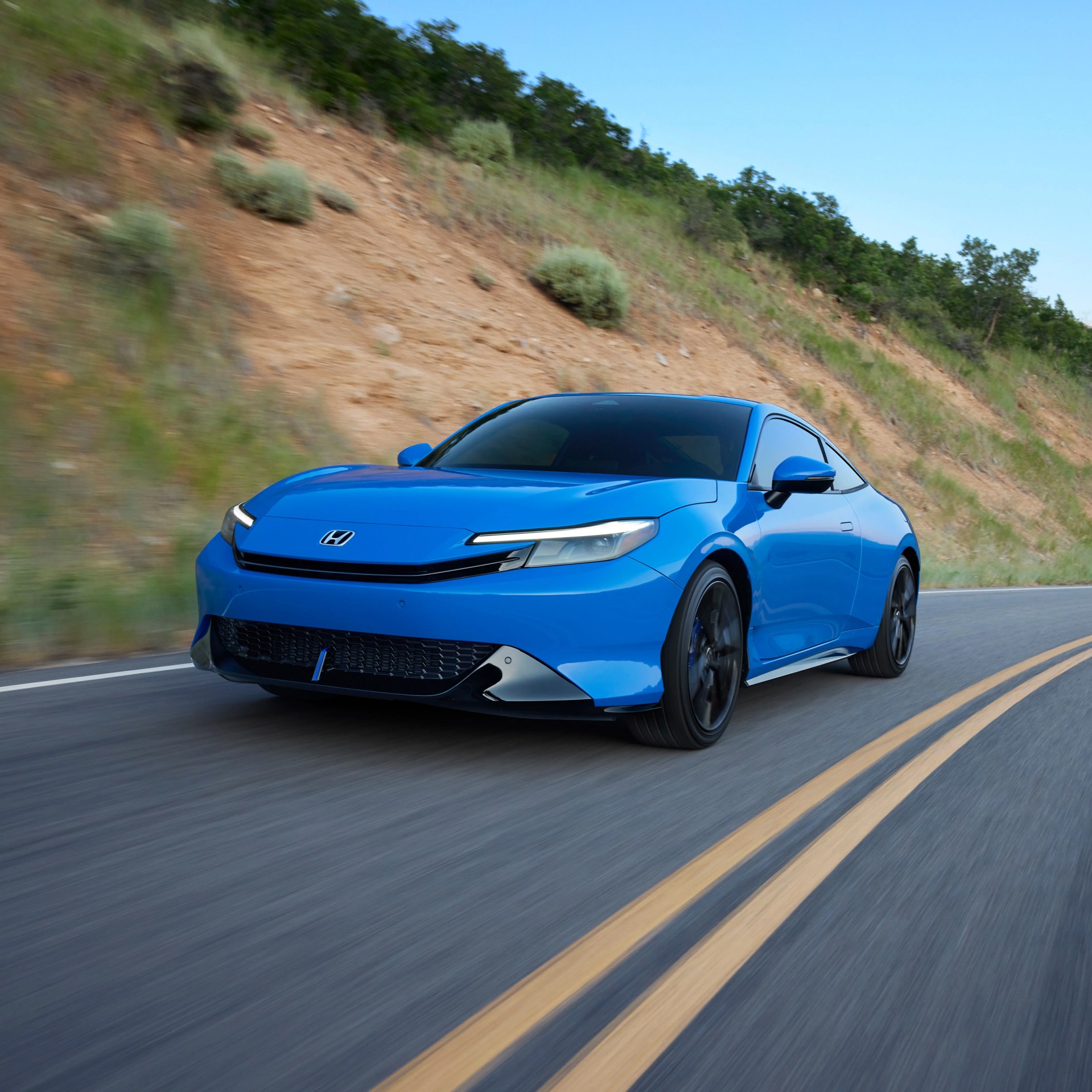 Blue Honda electric coupe driving on a winding road with a hillside covered in grass and shrubs in the background. The car features sleek, modern headlights and black wheels.