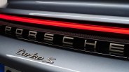 Close-up of the rear of a silver Porsche Turbo S, showing the illuminated red tail light strip, the "PORSCHE" lettering in metallic finish, and the "turbo S" badge below it.