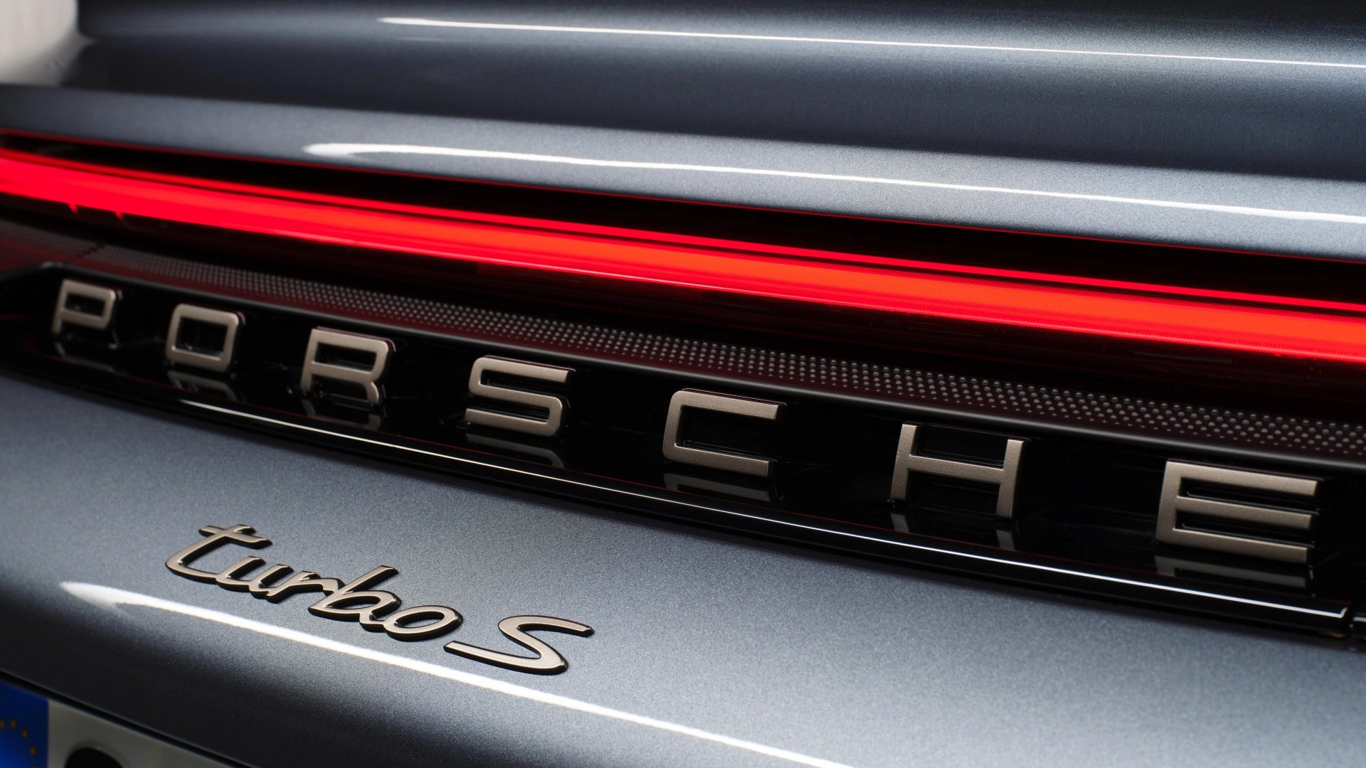 Close-up of the rear of a silver Porsche Turbo S, showing the illuminated red tail light strip, the "PORSCHE" lettering in metallic finish, and the "turbo S" badge below it.