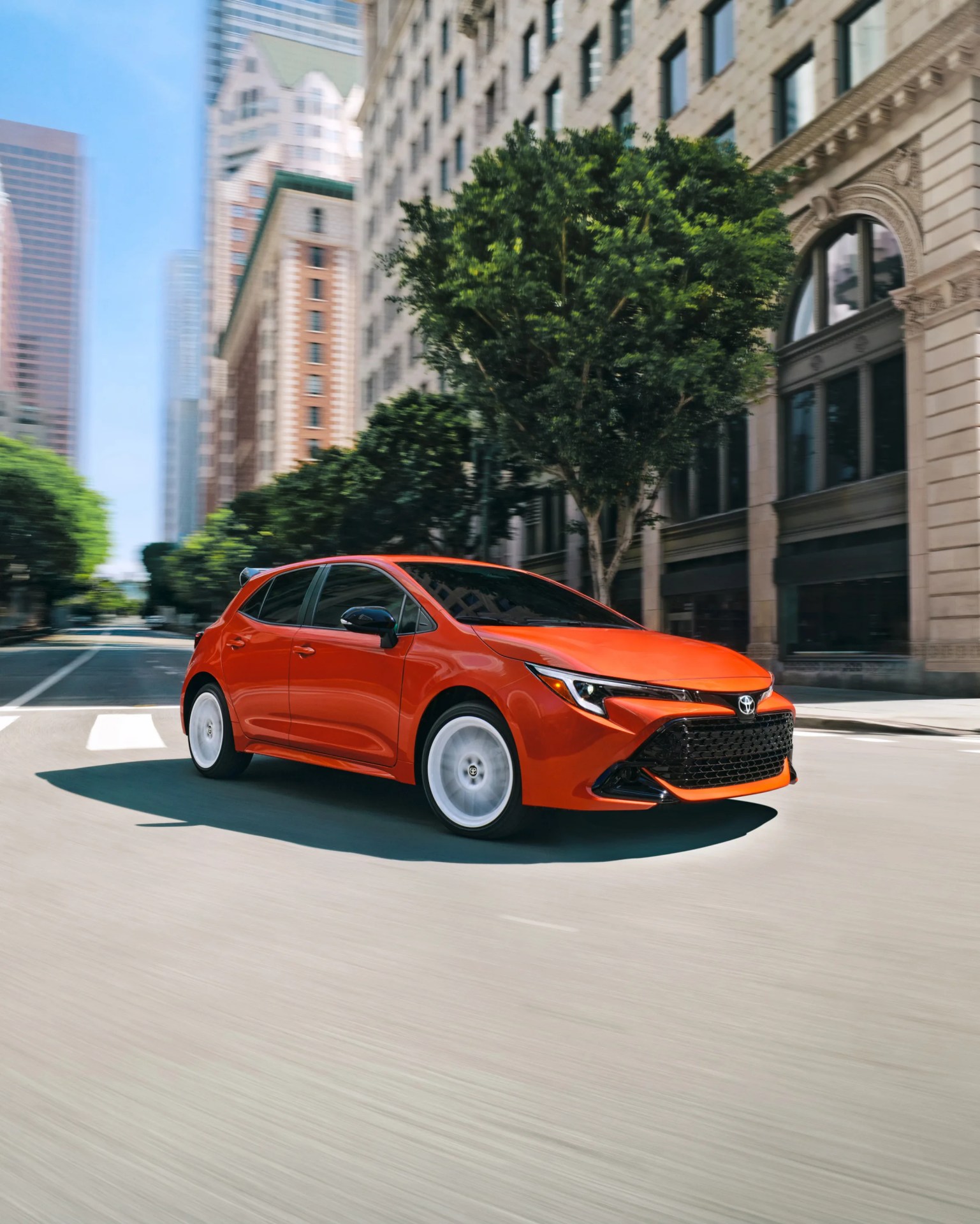 Orange compact hatchback car driving on a city street with tall buildings and trees in the background. The car has a sleek, modern design with black accents on the side mirrors and front grille.