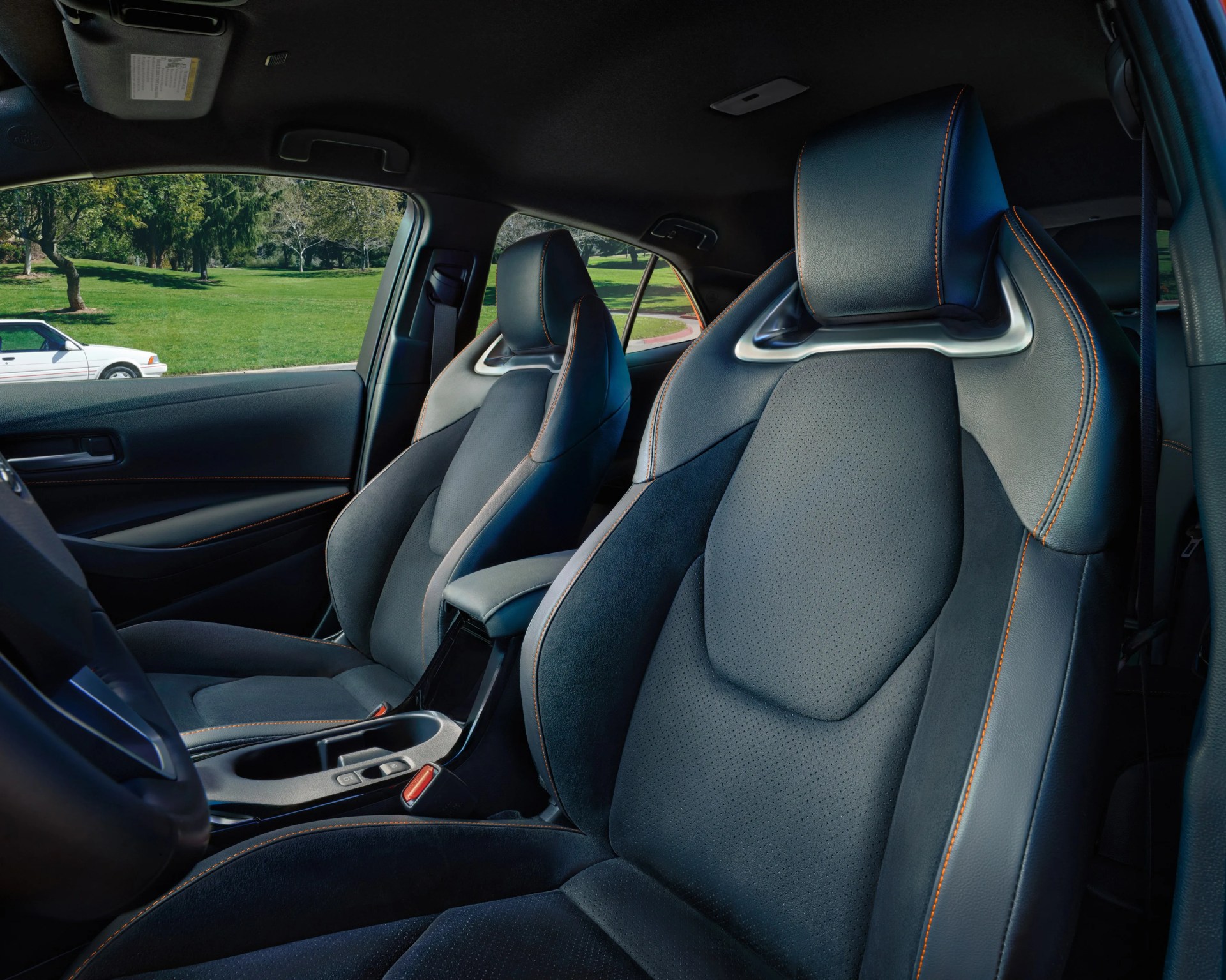 Two black leather and suede car seats with orange stitching are shown inside a vehicle. The seats have a sporty design with integrated headrests and metallic accents. The interior door panel and center console with cup holders and controls are also visible. Outside the window, there is a green grassy area with trees and a white car parked nearby.