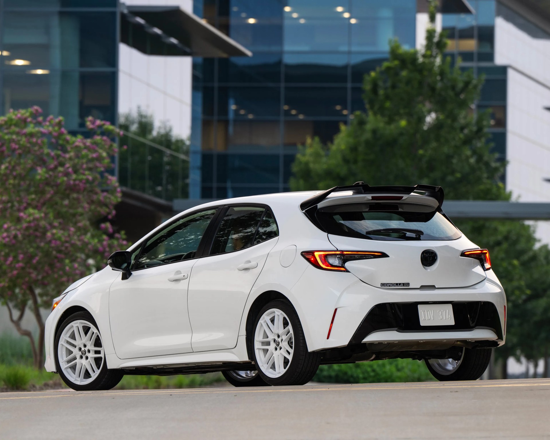 White Toyota Corolla hatchback with sporty rear spoiler and white wheels, parked on a street in an urban setting with modern buildings and greenery in the background.