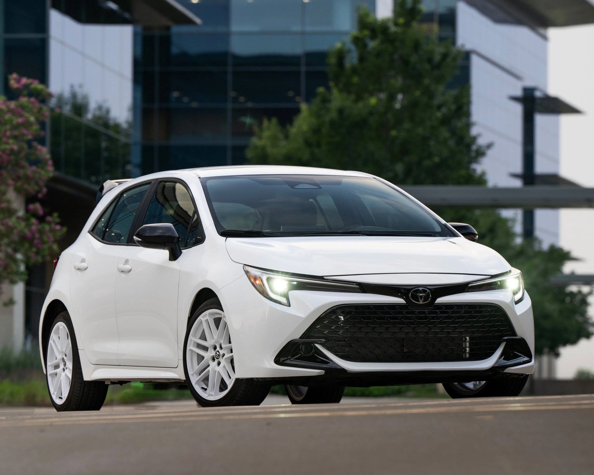 White Toyota hatchback with black accents and white wheels, parked on a street in front of modern glass buildings and greenery. The car's headlights are on, and it has a sporty, sleek design.