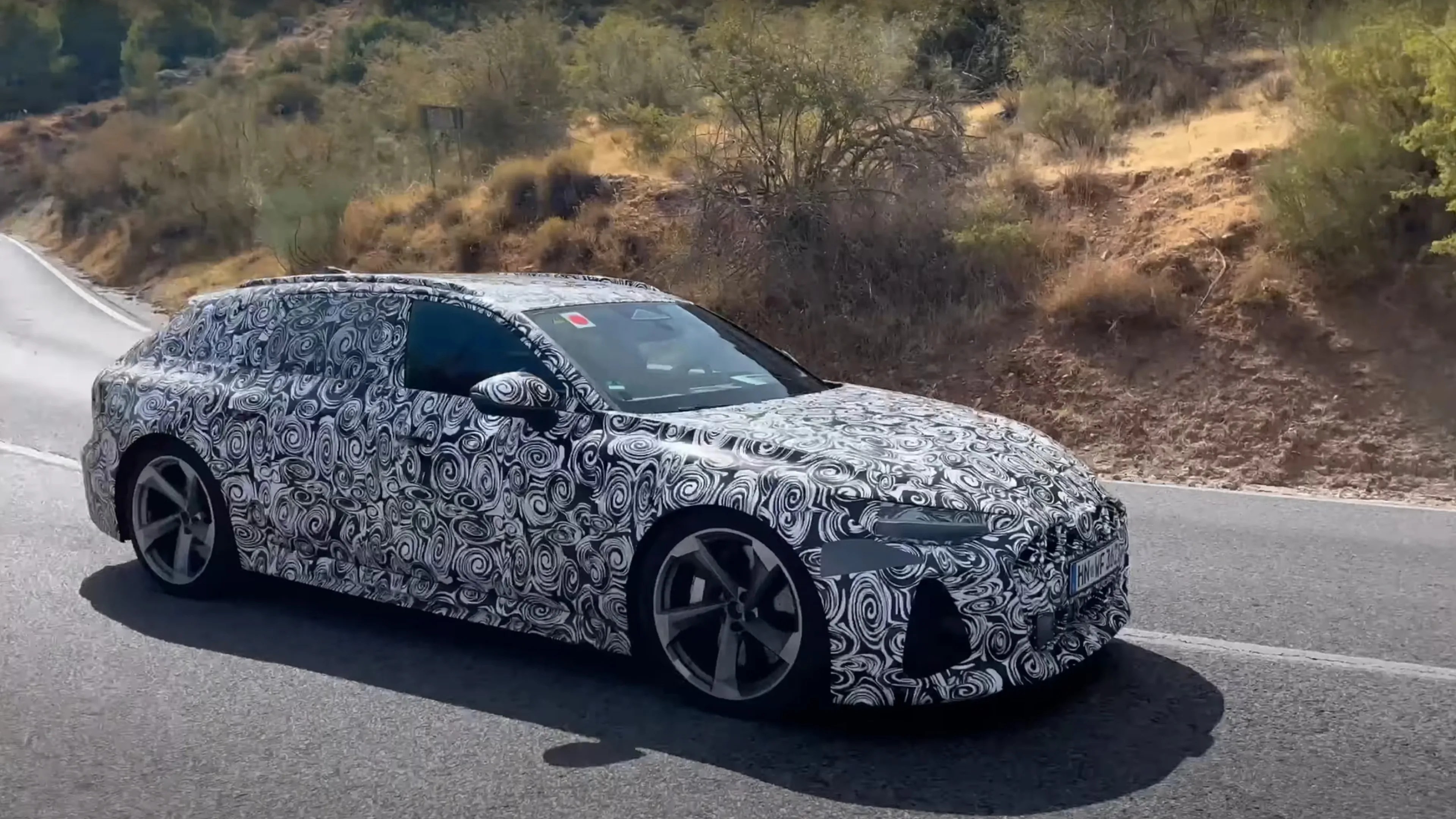 A camouflaged prototype car with a black and white swirl pattern covering its entire body, driving on a paved road in a dry, hilly area with sparse vegetation. The car has a sleek, sporty design with large alloy wheels and tinted windows.
